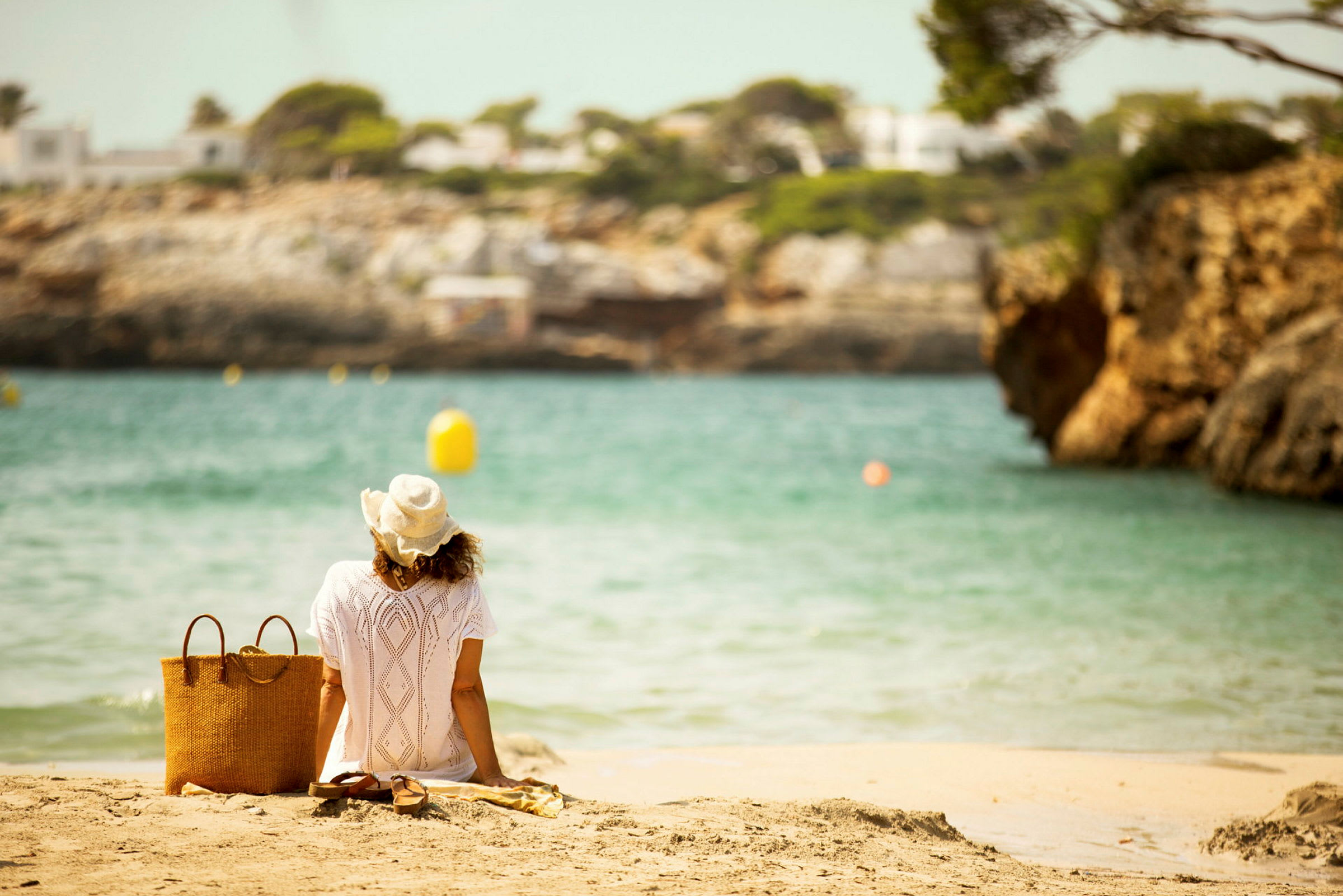 En person i vit klänning och hatt sitter på en strand med utsikt över vattnet. Bredvid personen finns en korgväska och ett par sandaler. (AI-genererad text)