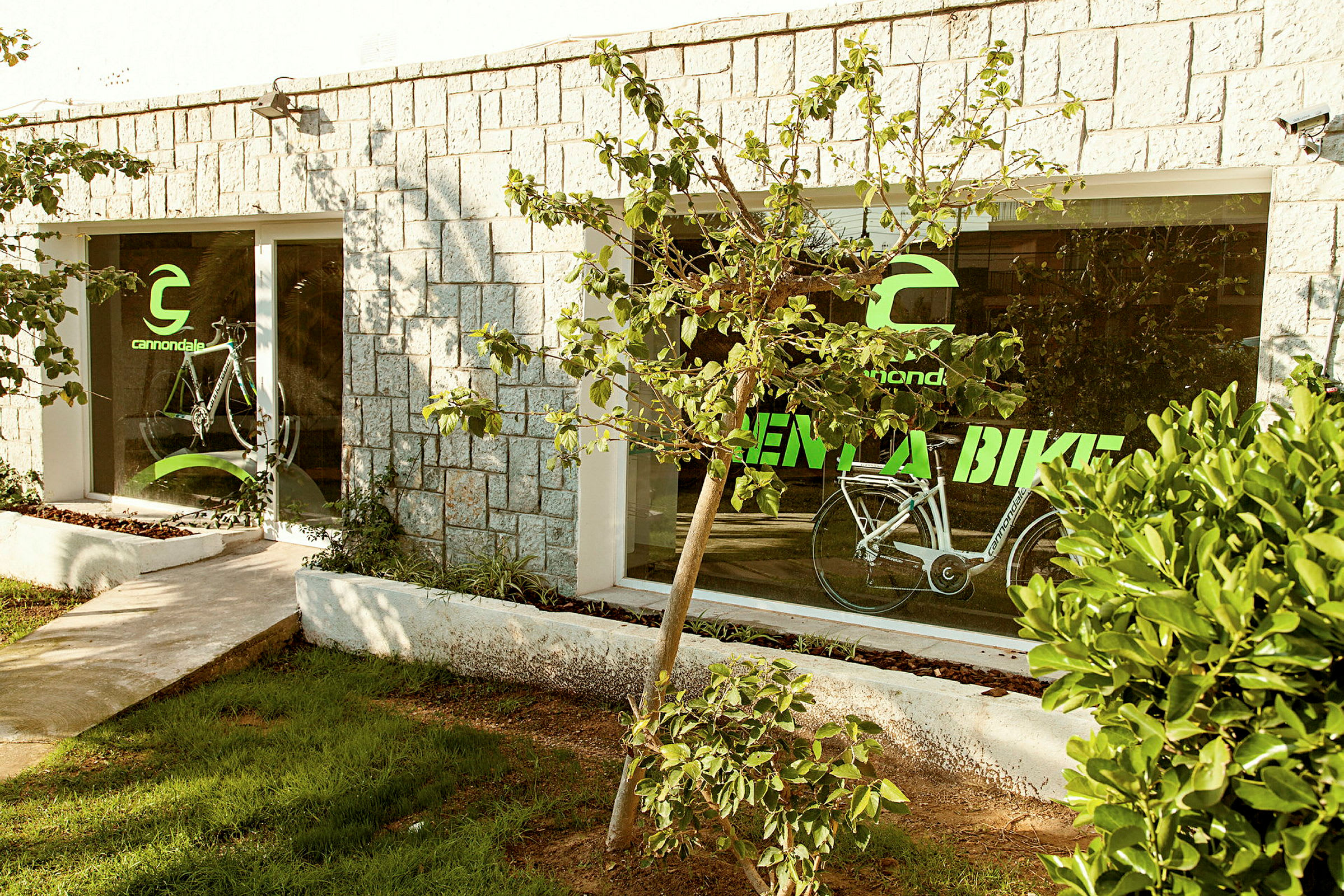 En byggnad med stenväggar och stora glasfönster. På fönstren finns texten "Cannondale Rent a Bike" och en bild av en cykel. Framför byggnaden växer träd och buskar. (AI-genererad text)