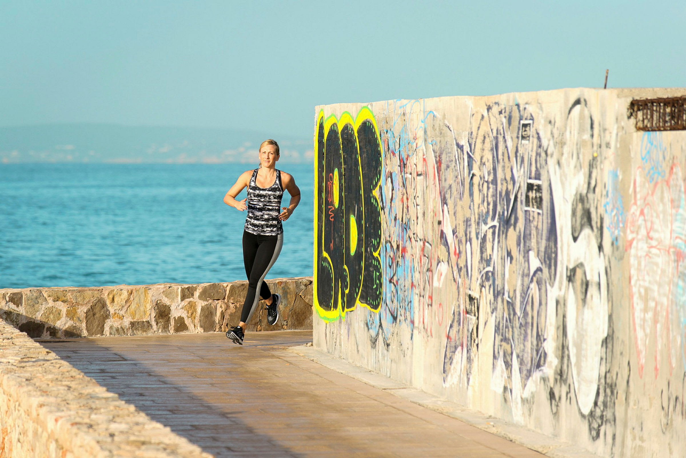 Person joggar längs en strandpromenad med havet i bakgrunden och en vägg målad med graffiti. (AI-genererad text)
