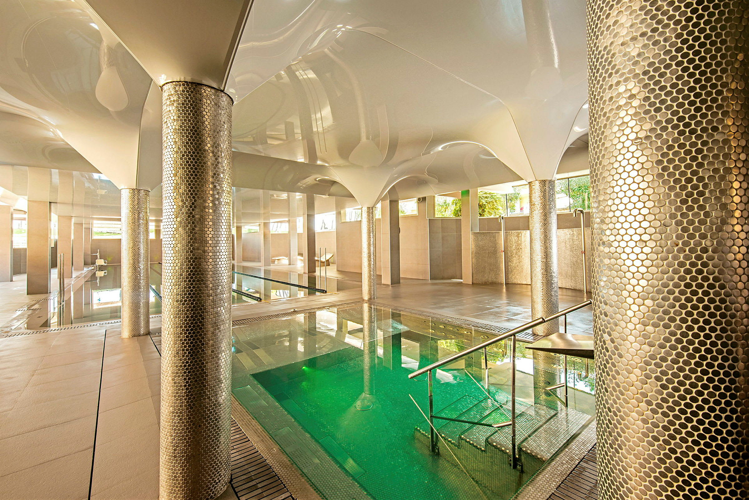 Ett modernt inomhus spaområde med en grönskimrande pool omgiven av dekorativa metallpelare och vitt tak. (AI-genererad text)