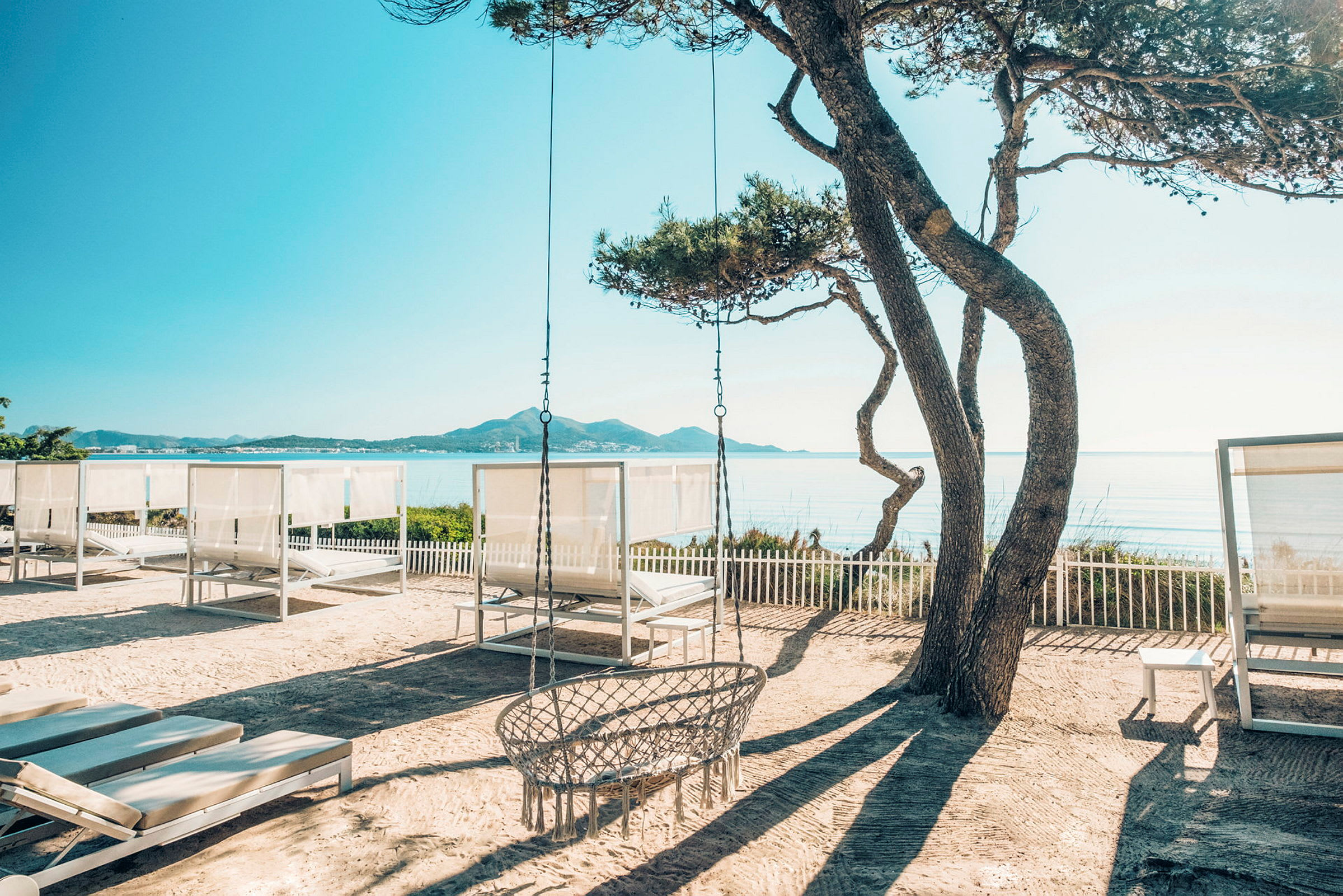 En strand med solsängar och träd i förgrunden, med havet och berg i bakgrunden. (AI-genererad text)