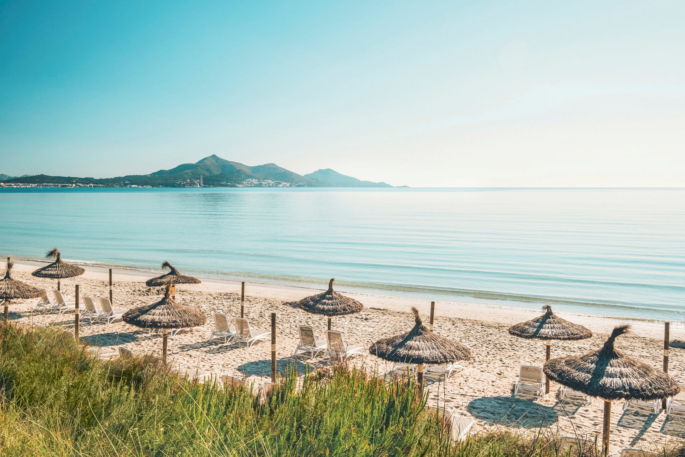 En sandstrand med solstolar och halmtakparaplyn med havet och berg i bakgrunden. (AI-genererad text)