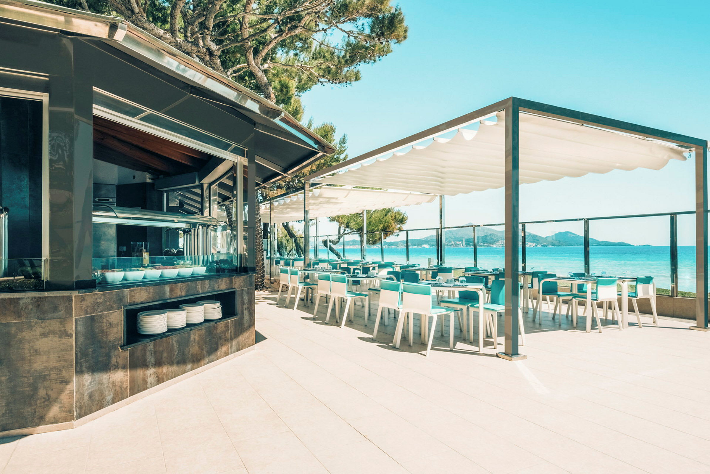 En bild visar en utomhusrestaurang med bord och stolar i vitt och turkost, nära havet och under en soltak. (AI-genererad text)