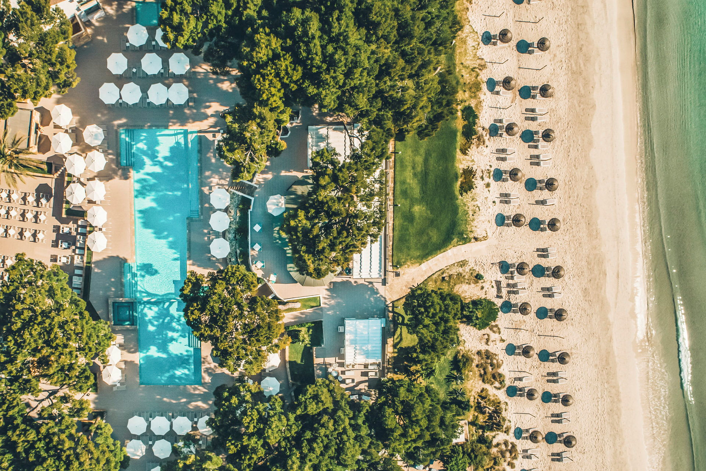 Flygfoto av en strand med solstolar och parasoller, en pool omgiven av grönska och byggnader. (AI-genererad text)
