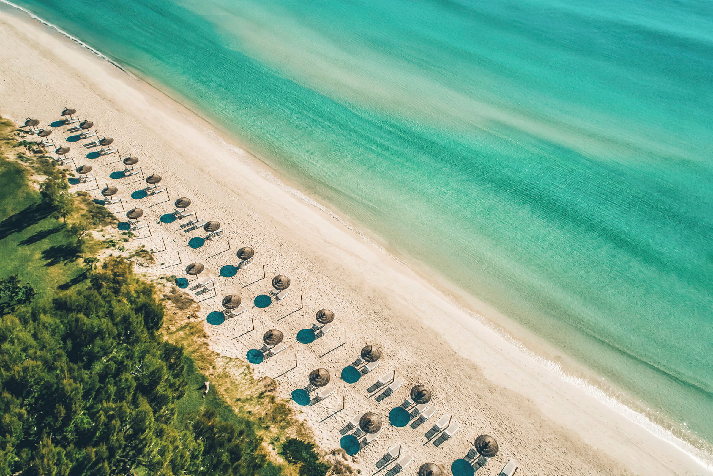 En flygbild av en sandstrand med parasoller och solstolar intill det turkosa havet. (AI-genererad text)