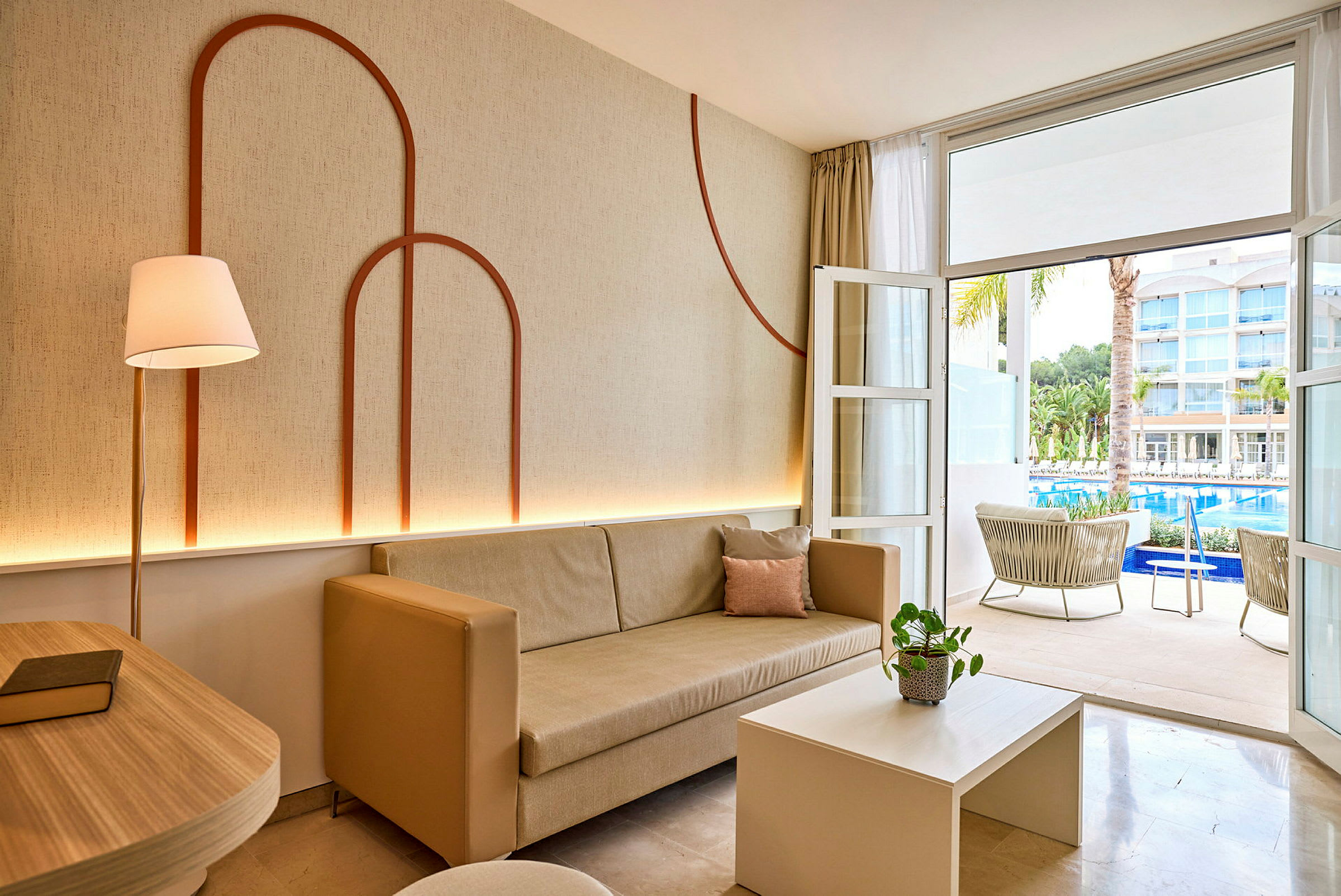 Vardagsrum med beige soffa och dekorerad vägg, öppning mot terrass med utsikt över pool och hotellbyggnader. (AI-genererad text)