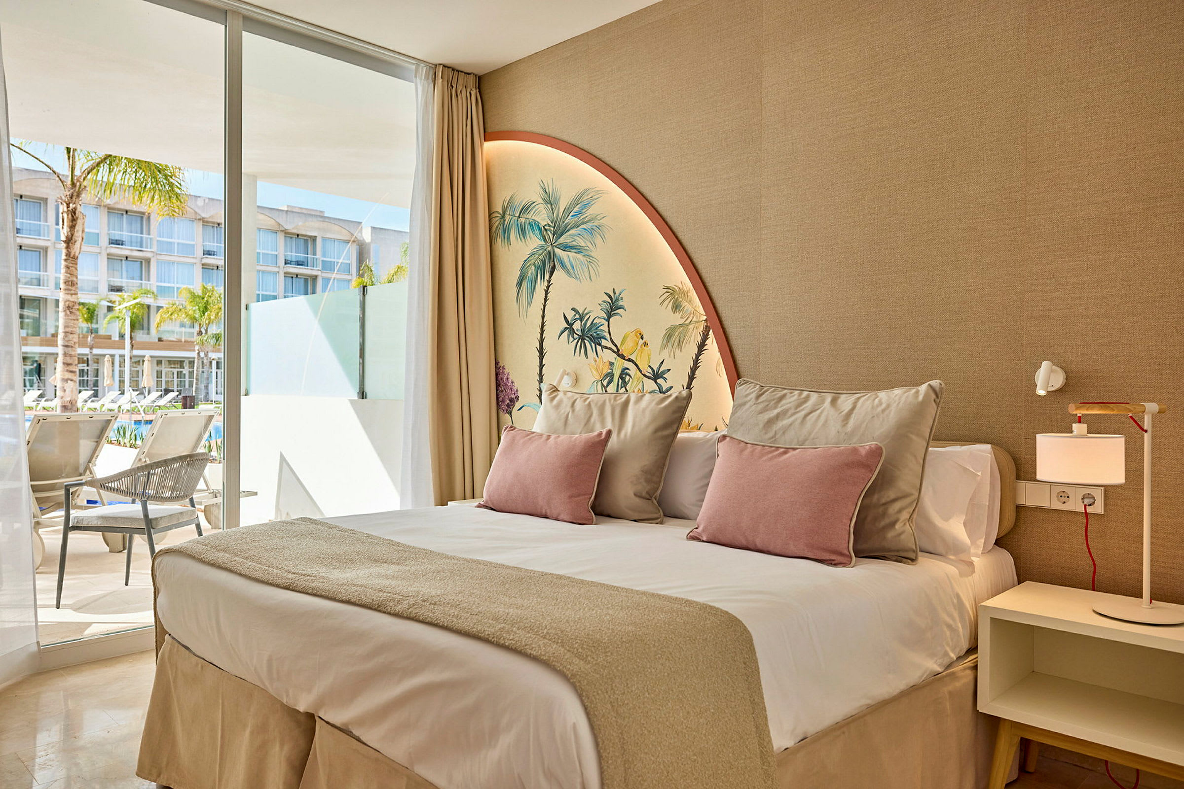 Ett hotellrum med en dubbelsäng, dekorerat med ljusa kuddar och en väggmålning som visar palmer. Fönster med utsikt över en utomhuspoolområde. (AI-genererad text)