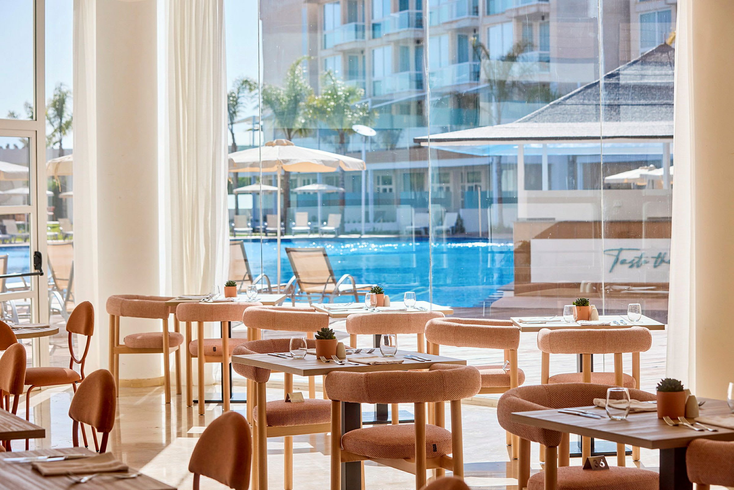 Interiör av en restaurang med bord och stolar, stora fönster med utsikt över en pool och byggnader. (AI-genererad text)