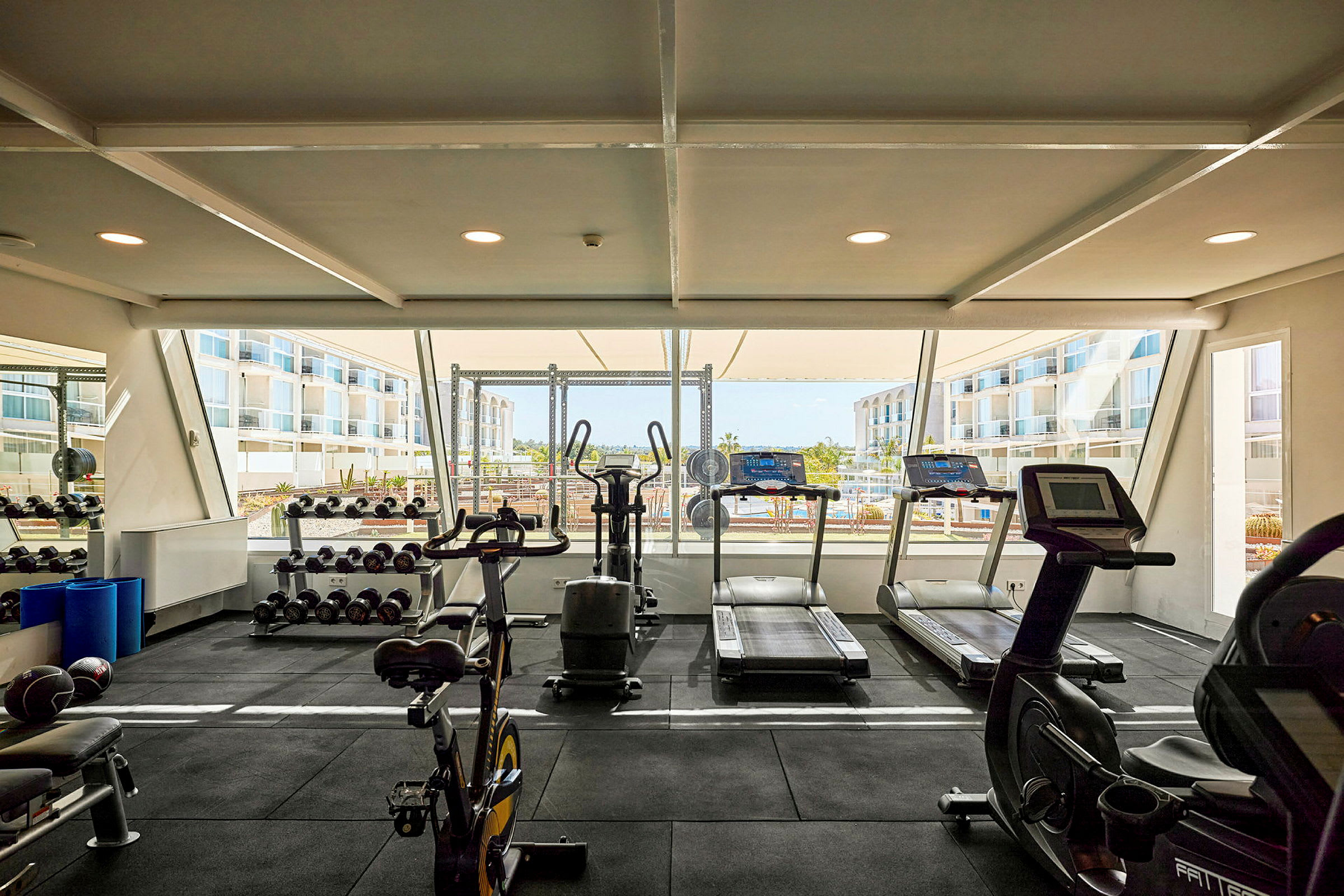 Gym med träningsmaskiner och vikter samt stora fönster med utsikt mot omgivande byggnader. (AI-genererad text)