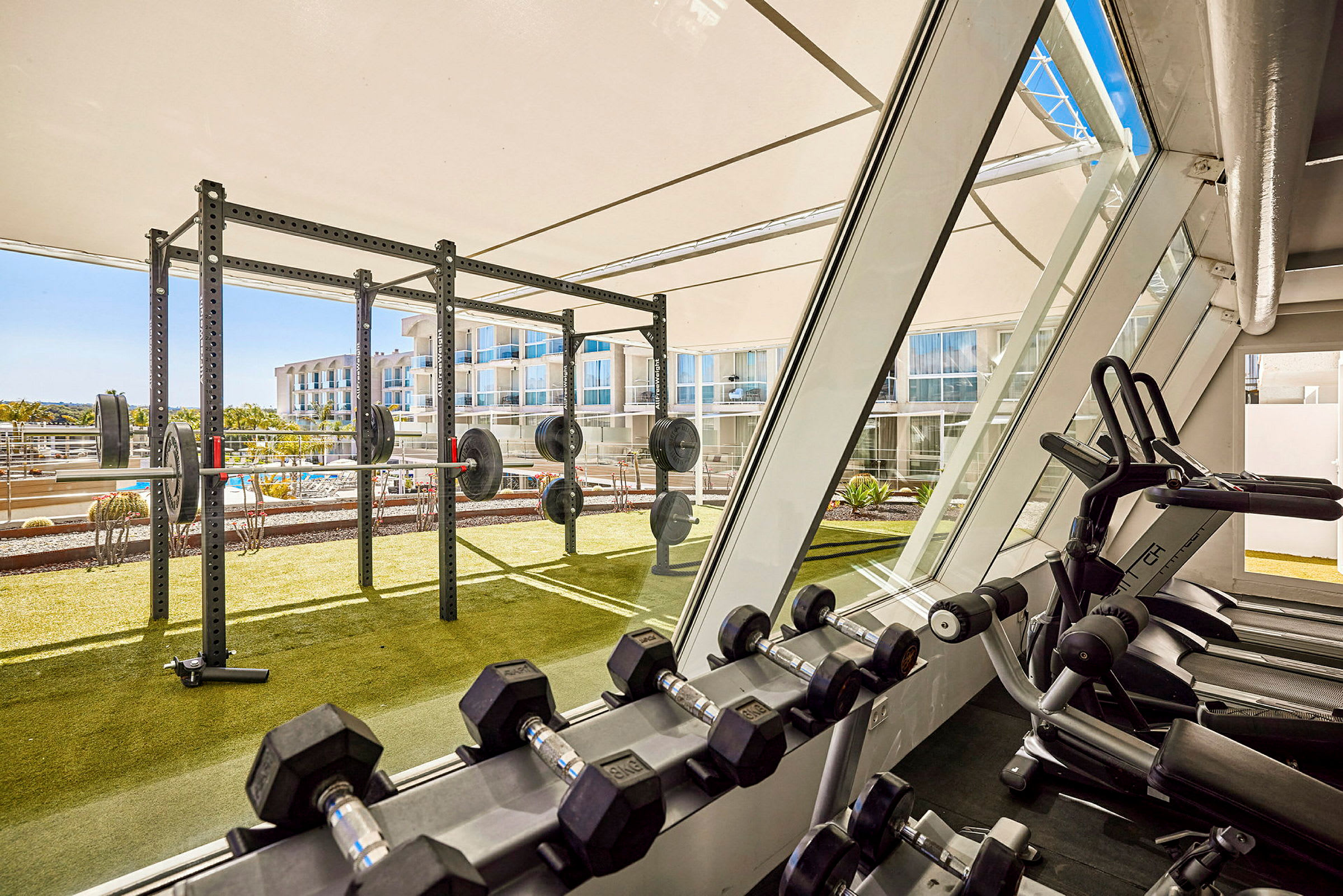 Ett gym med träningsutrustning som hantlar och styrkelyftställ, med utsikt över utomhusområdet. (AI-genererad text)