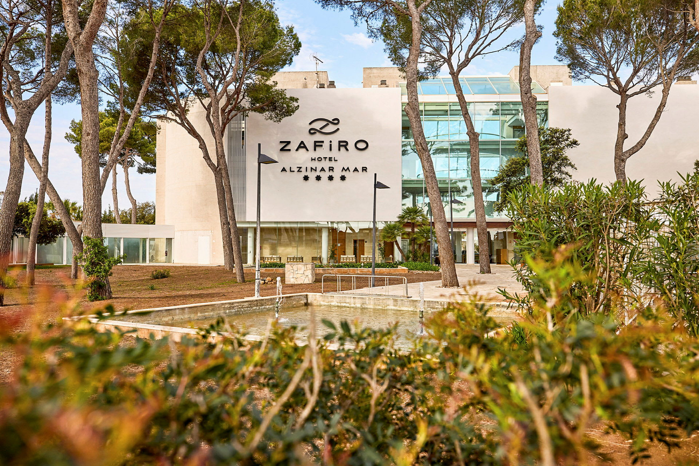 Hotellbyggnaden Zafiro Alzinar Mar med träd och en fontän i förgrunden. (AI-genererad text)