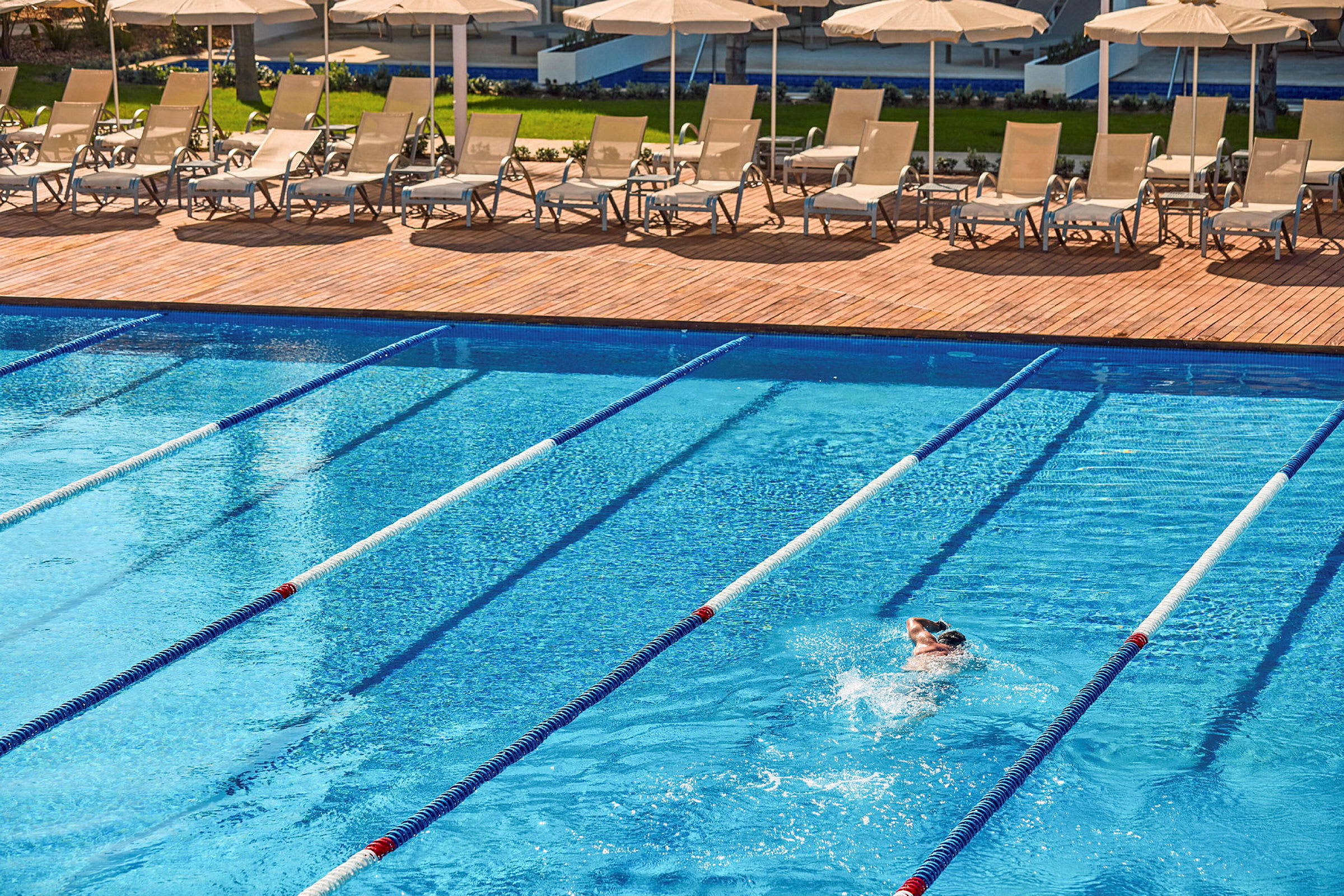 En person simmar i en pool med markerade banor. Solstolar och parasoller syns på terrassen i bakgrunden. (AI-genererad text)