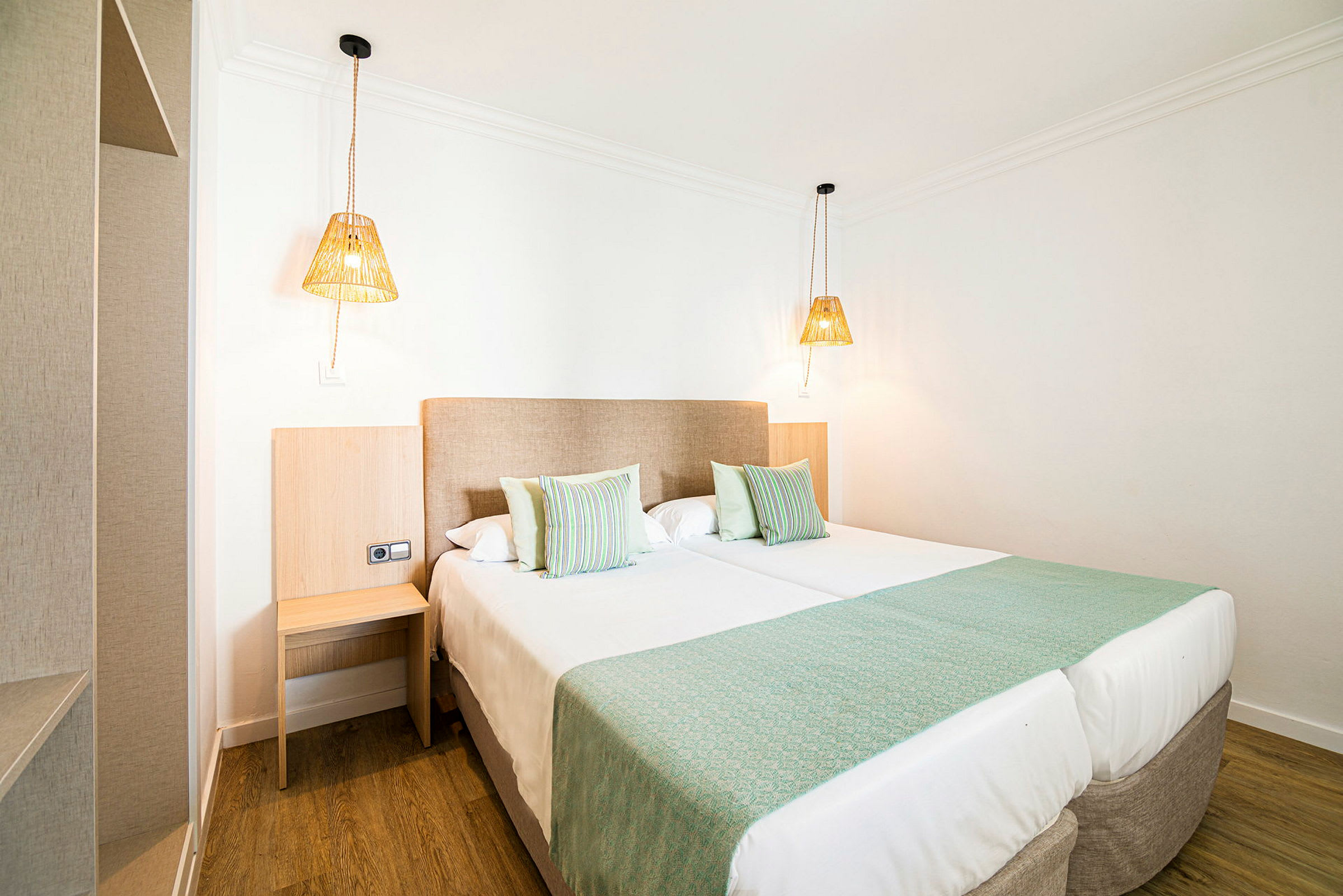 Ett hotellrum med två bäddade enkelsängar, gröna sängöverkast och kuddar samt två hängande lampor. (AI-genererad text)
