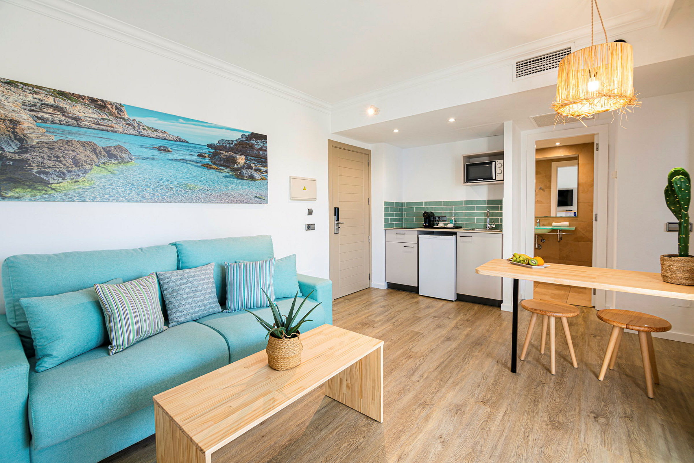 Ett hotellrum med ljusblå soffa och en modern kokvrå. Väggdekoration föreställande en strand. (AI-genererad text)