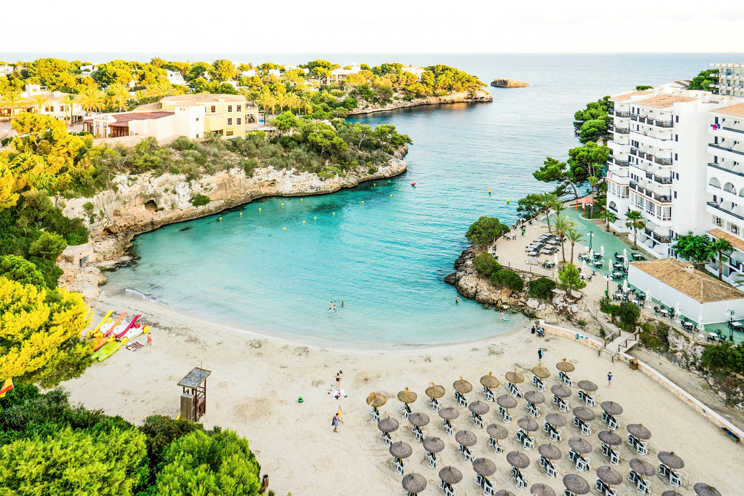 Strand med parasoller och solstolar vid en turkos vik omgiven av klippor och grönska. (AI-genererad text)