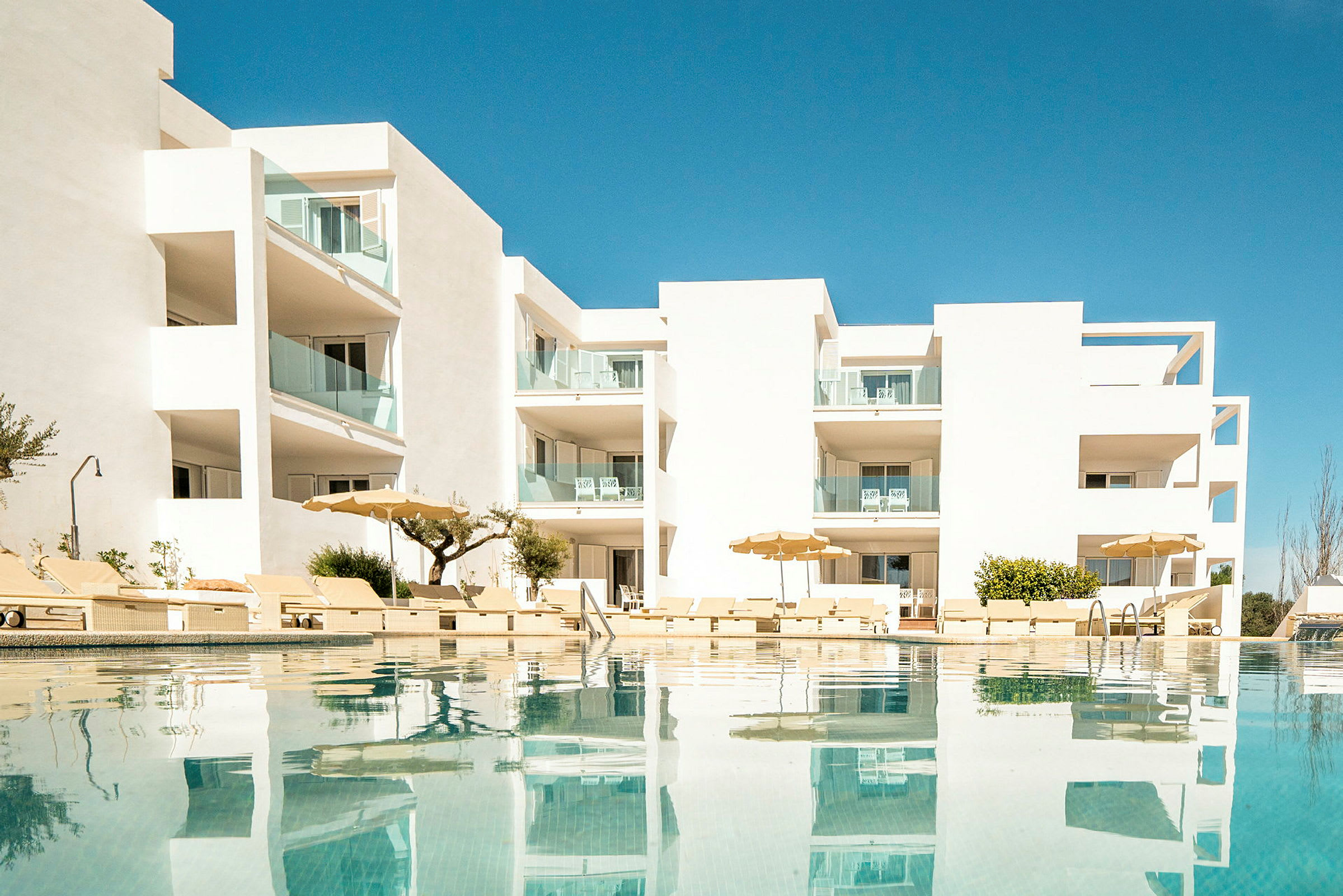 En modern vit byggnad med pool och solstolar framför, reflekterad i vattnet under en klarblå himmel. (AI-genererad text)