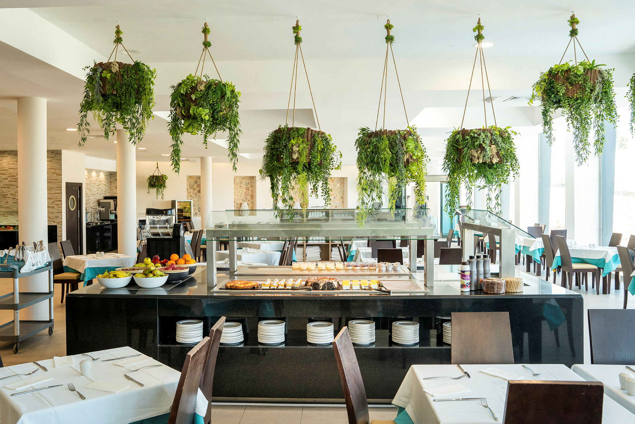 Restaurang med bord, stolar och buffé samt hängande gröna växter. (AI-genererad text)