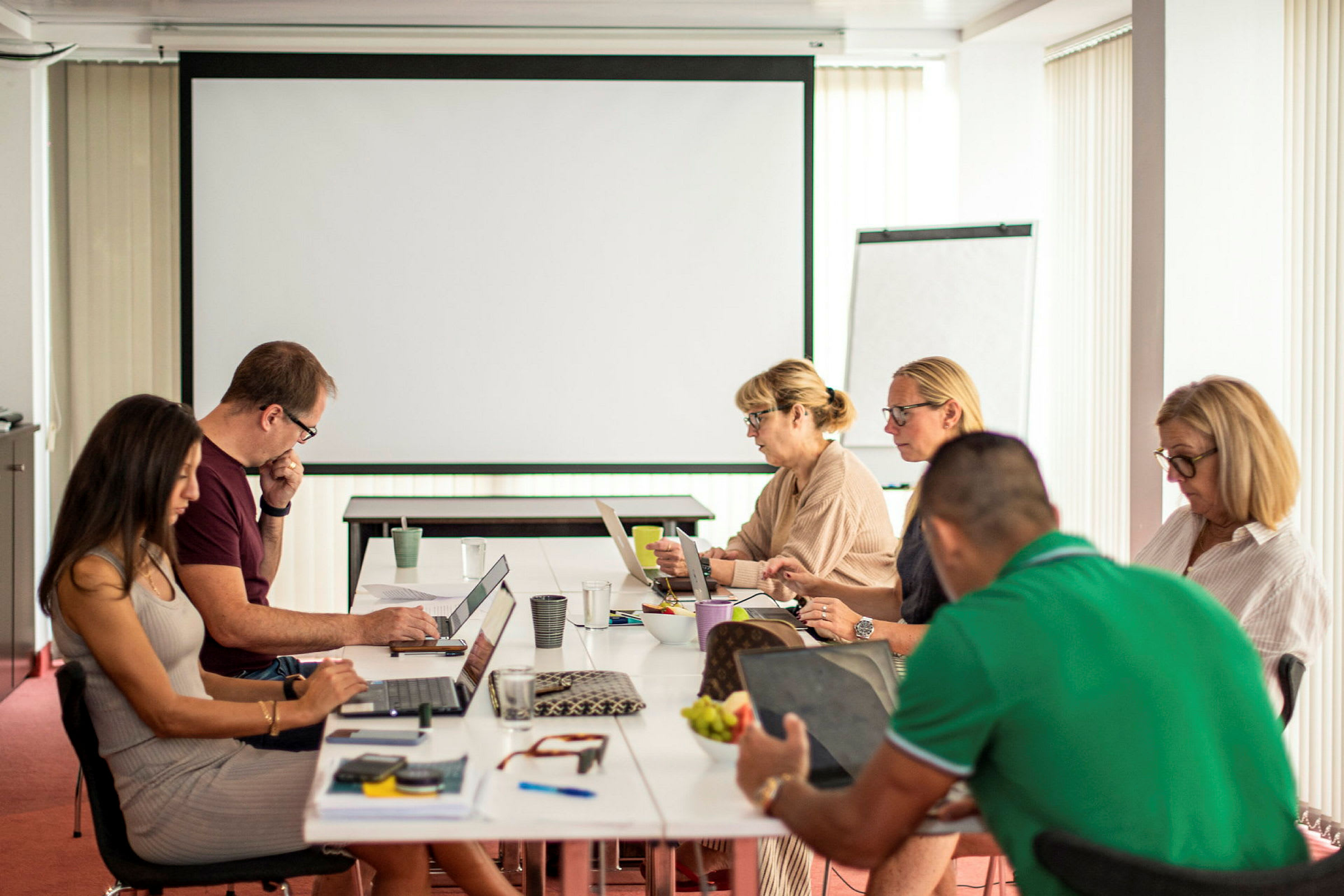 En grupp människor sitter runt ett bord med laptops och anteckningar i ett mötesrum. (AI-genererad text)