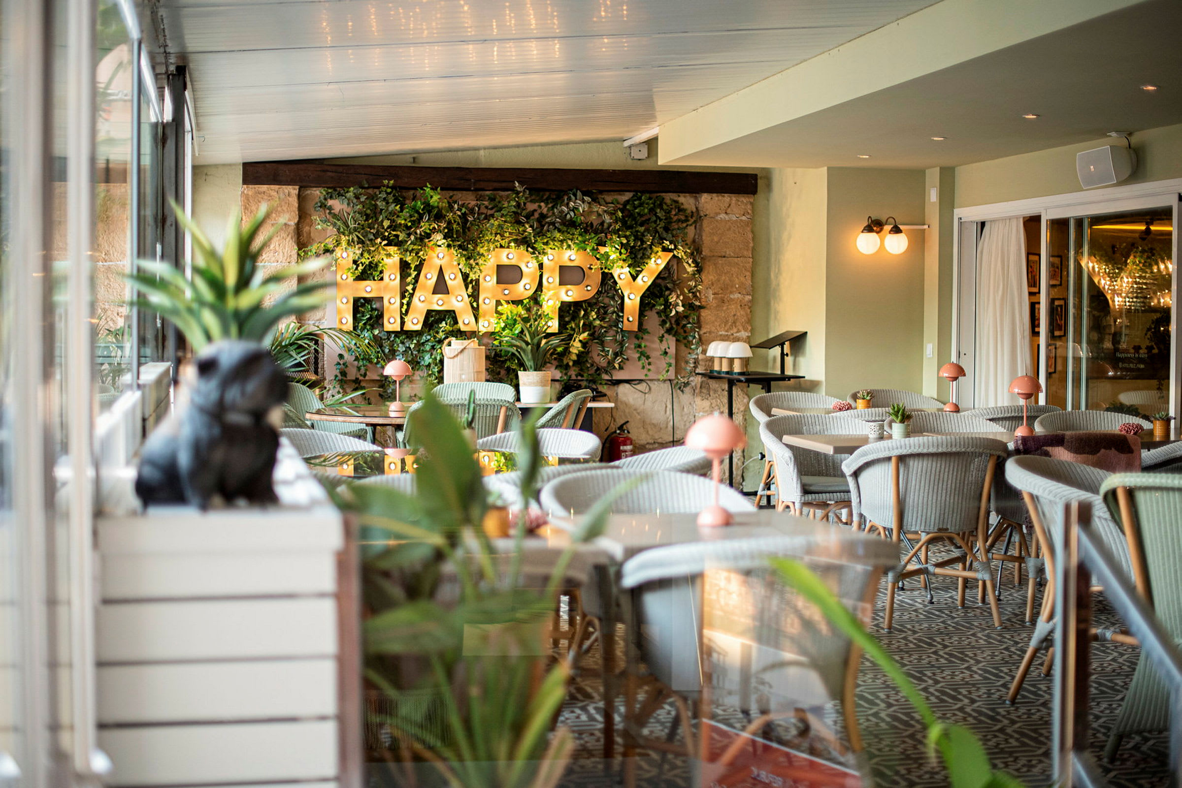 Interiör av en restaurang eller cafémiljö med gröna växter, belysning och texten 'HAPPY' på väggen. (AI-genererad text)