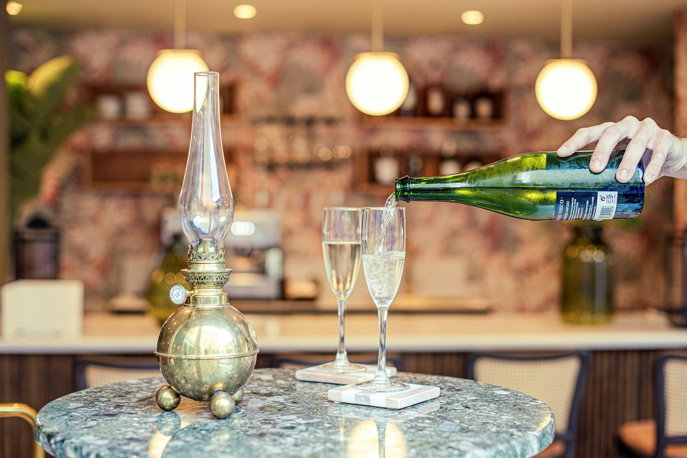 En antik oljelampa på ett bord bredvid två champagneglas. En hand häller dryck i ett av glasen. (AI-genererad text)