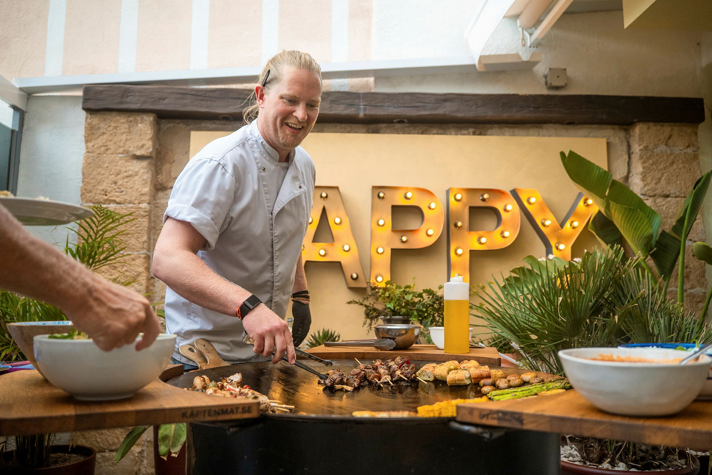 Person tillagar mat på en grill med ordet 'Happy' upplyst i bakgrunden. (AI-genererad text)