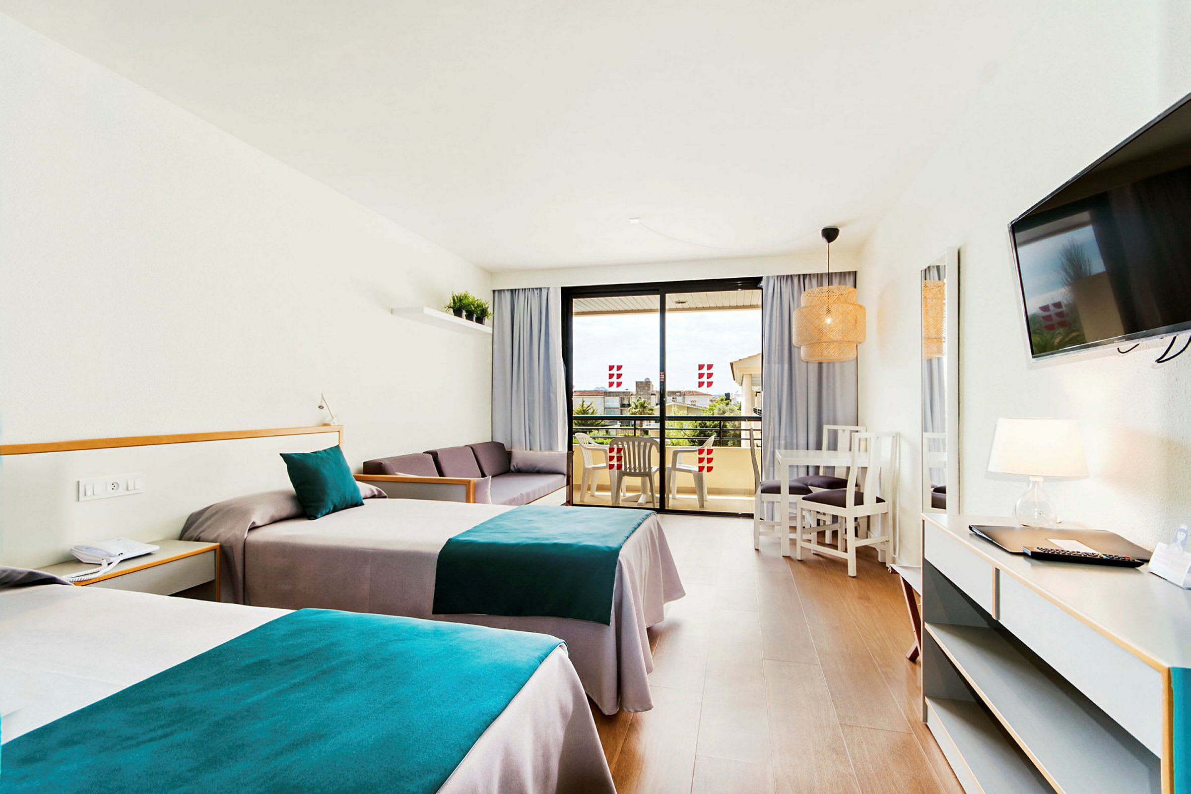 Ett hotellrum med två enkelsängar med blå och grå sängkläder, en soffa, ett bord med stolar och en balkong i bakgrunden. (AI-genererad text)