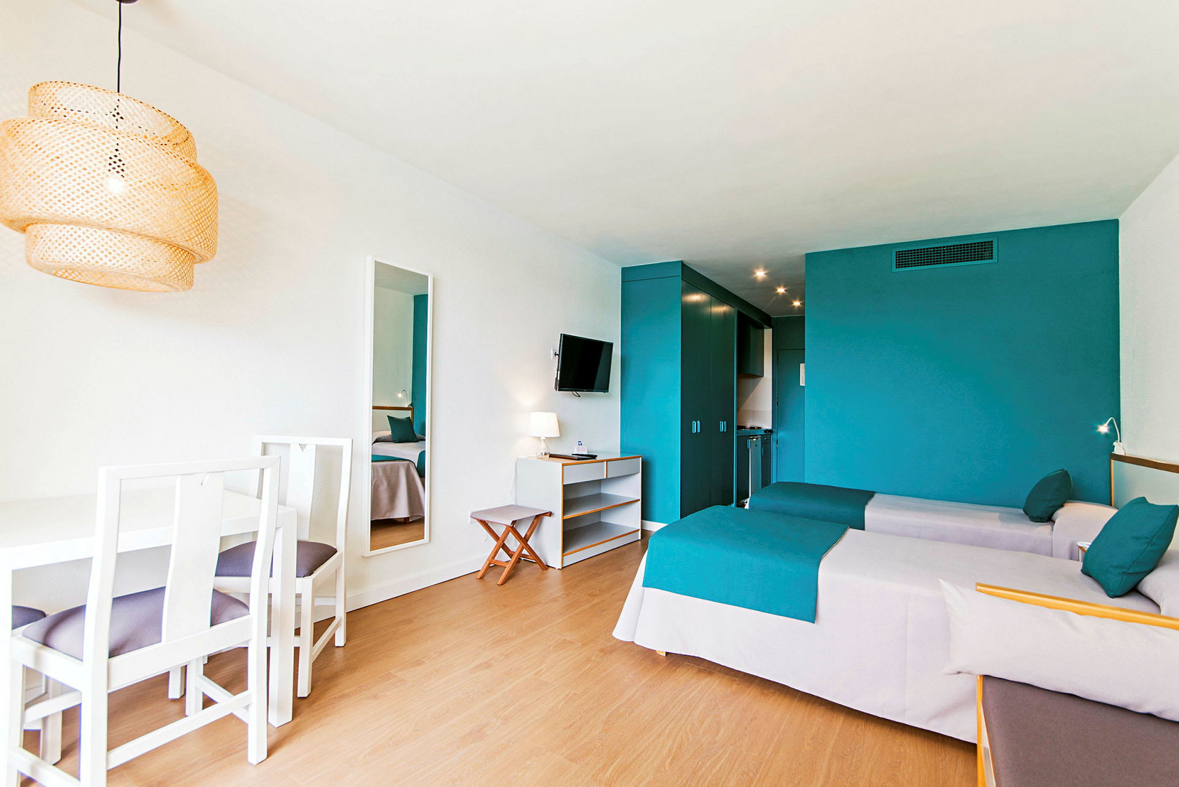 Ett hotellrum med två enkelsängar, turkosa sängöverkast och en vit matplats med fyra stolar. En vägg har en spegel och en monterad TV. (AI-genererad text)