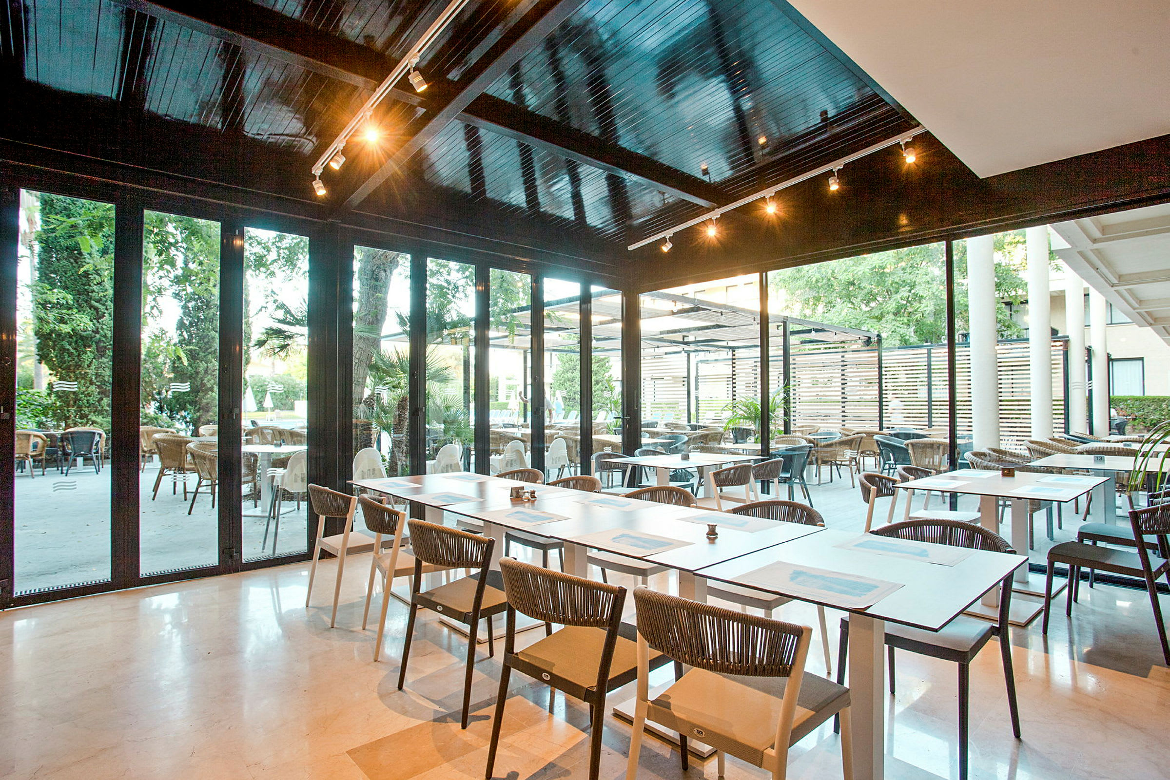 En modern restaurang med sittplatser både inomhus och utomhus. Bord och stolar är placerade på ett ljust golv, med glasväggar som ger utsikt över uteområdet. (AI-genererad text)