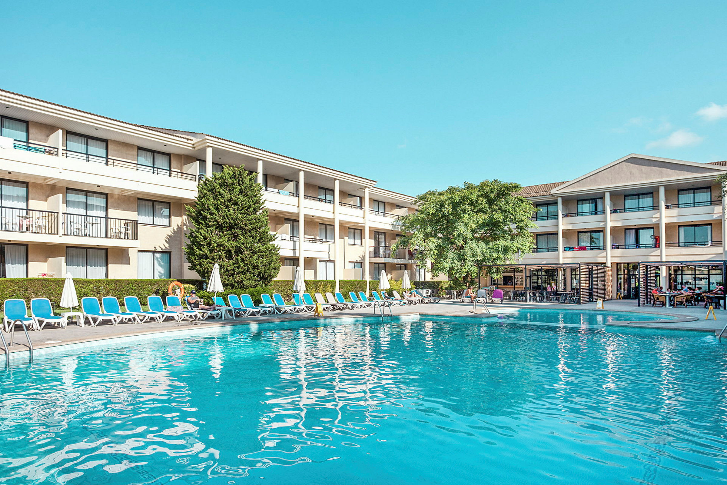 Bild som visar ett hotell med tre våningar, solstolar och parasoller runt en swimmingpool. Vid kanten finns en grön växt och träd. (AI-genererad text)