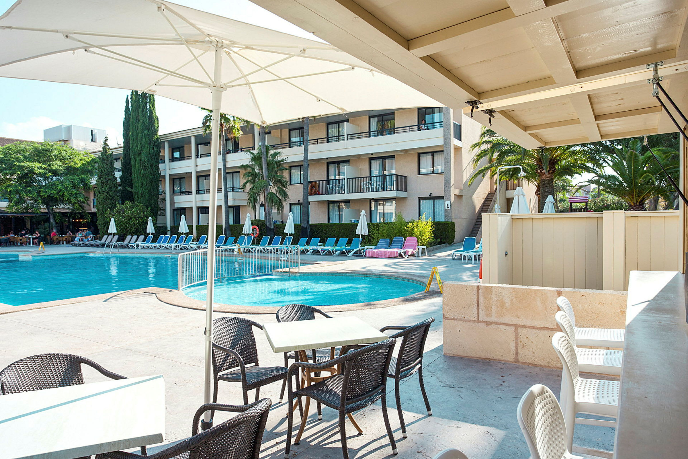 Poolområde med solstolar och parasoll vid en hotellbyggnad. I förgrunden finns bord och stolar. (AI-genererad text)