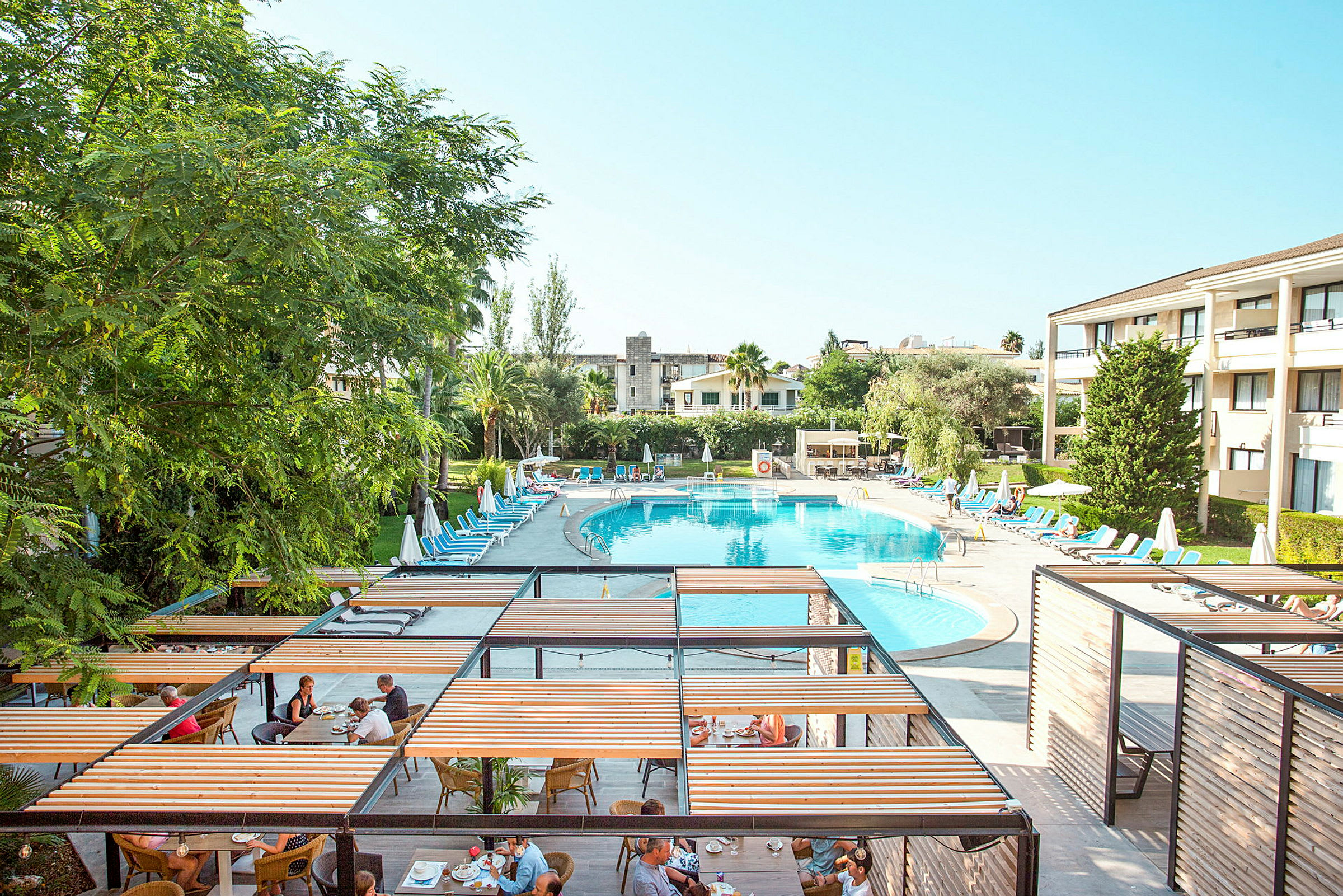 En semesteranläggning med en stor oval pool omgiven av solstolar och parasoller, samt en matplats med pergolor. (AI-genererad text)
