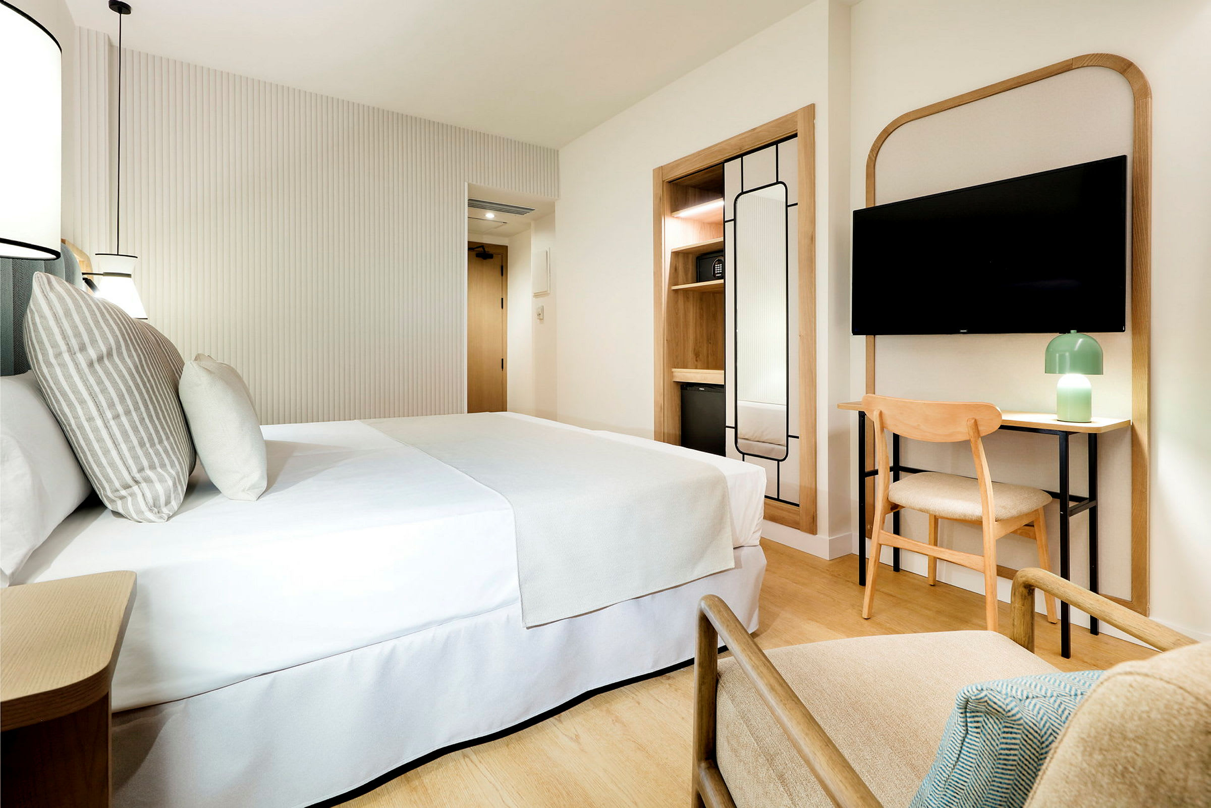 Ett modernt och ljust hotellrum med en dubbelsäng, en TV på väggen, en fåtölj, ett skrivbord med stol, och en minimalistisk inredning. (AI-genererad text)