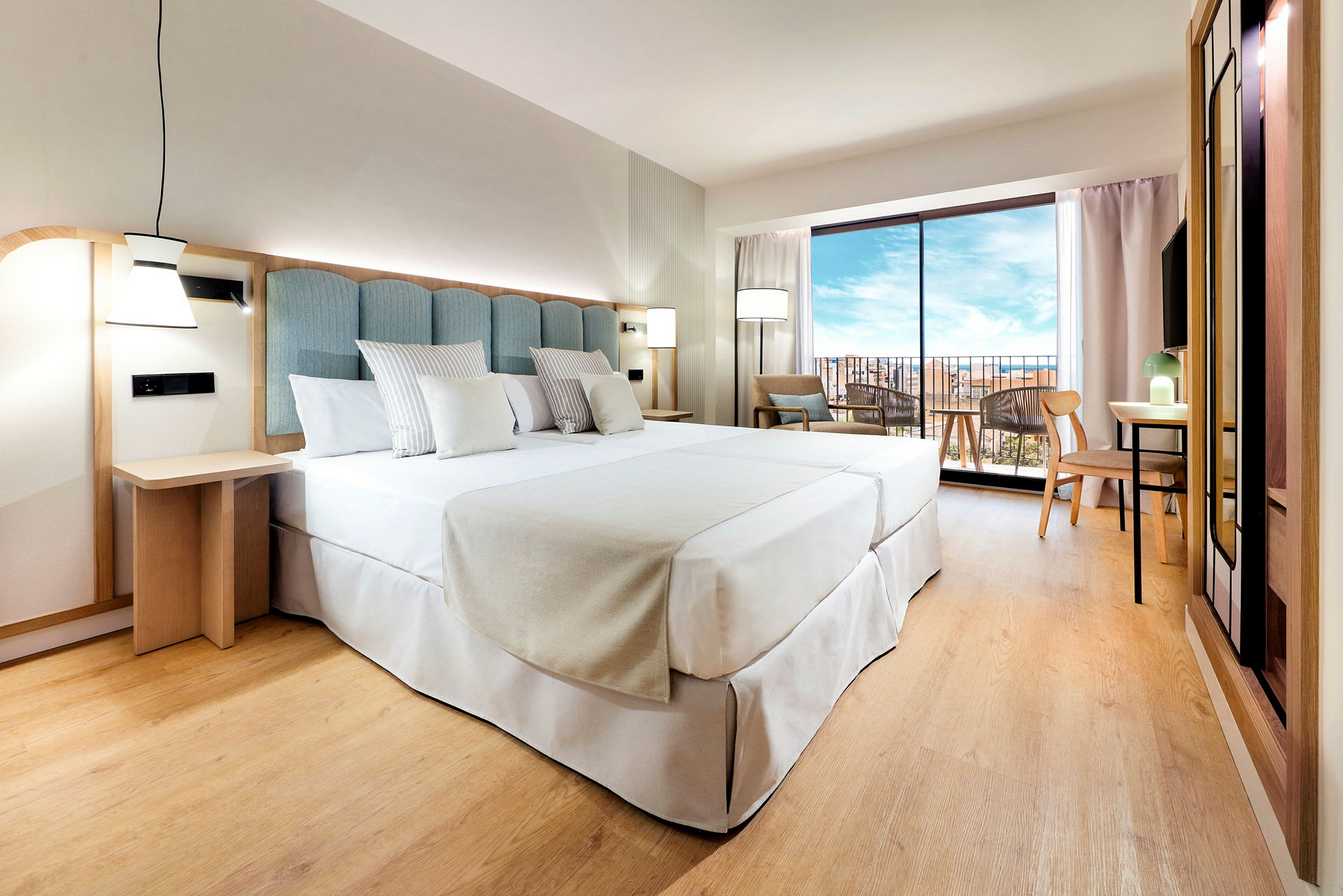 Ett hotellrum med en stor bäddad säng, paneler i ljust trä, och ett stort fönster som visar en vy utomhus. (AI-genererad text)