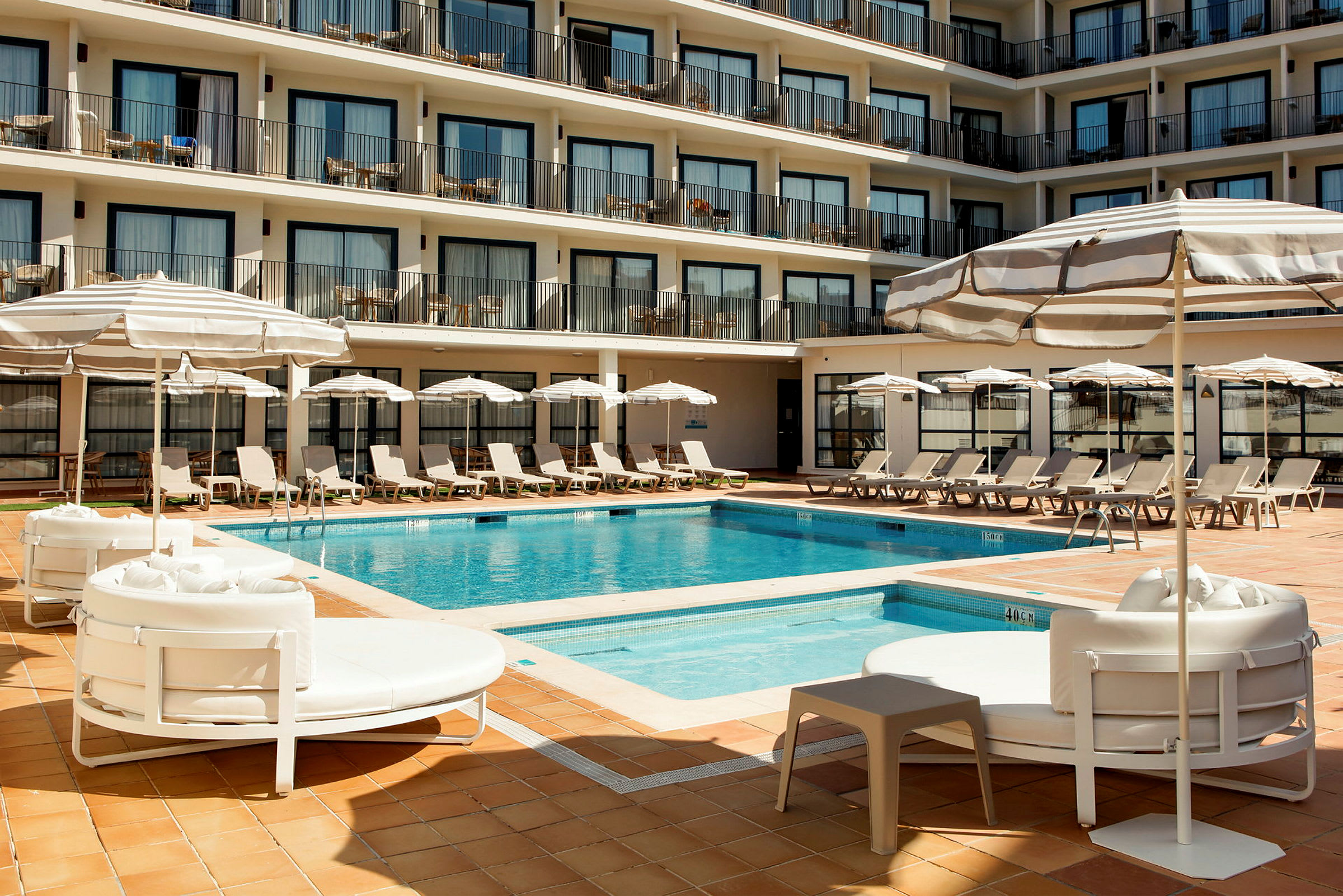 Ett hotellpoolområde med solstolar, parasoller och en pool omgiven av flera våningar med balkonger. (AI-genererad text)