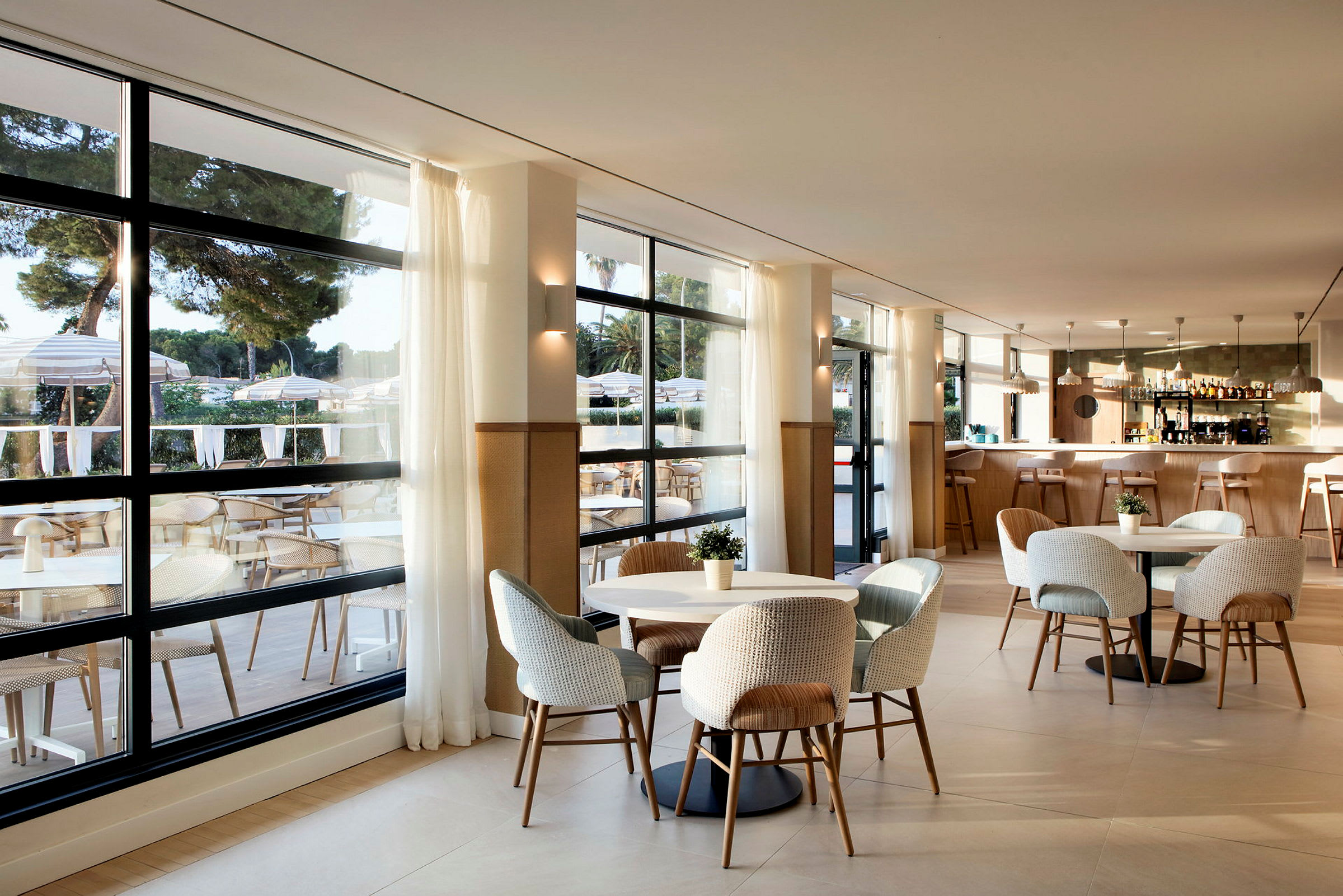 En modern restaurang med stora fönster, ljusa möbler och en välkomnande inredning. Utsikt mot en terrass med bord och parasoller. (AI-genererad text)