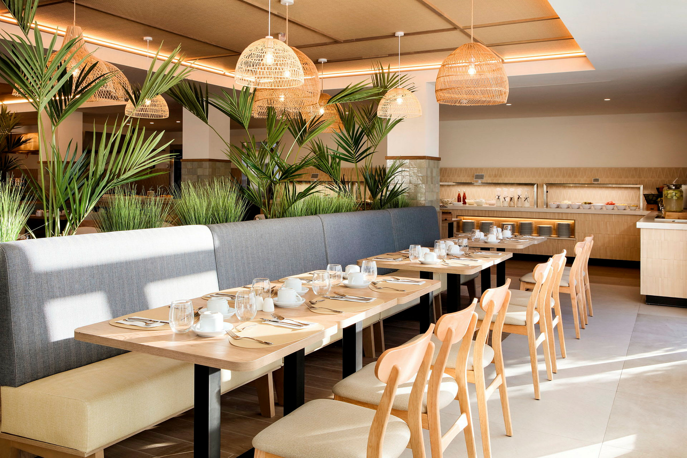 En ljus och modern restaurang med träbord, stolar och bänk av tyg. Dekorerad med gröna växter och flätade taklampor. Porcelin och glas dukat på borden. (AI-genererad text)