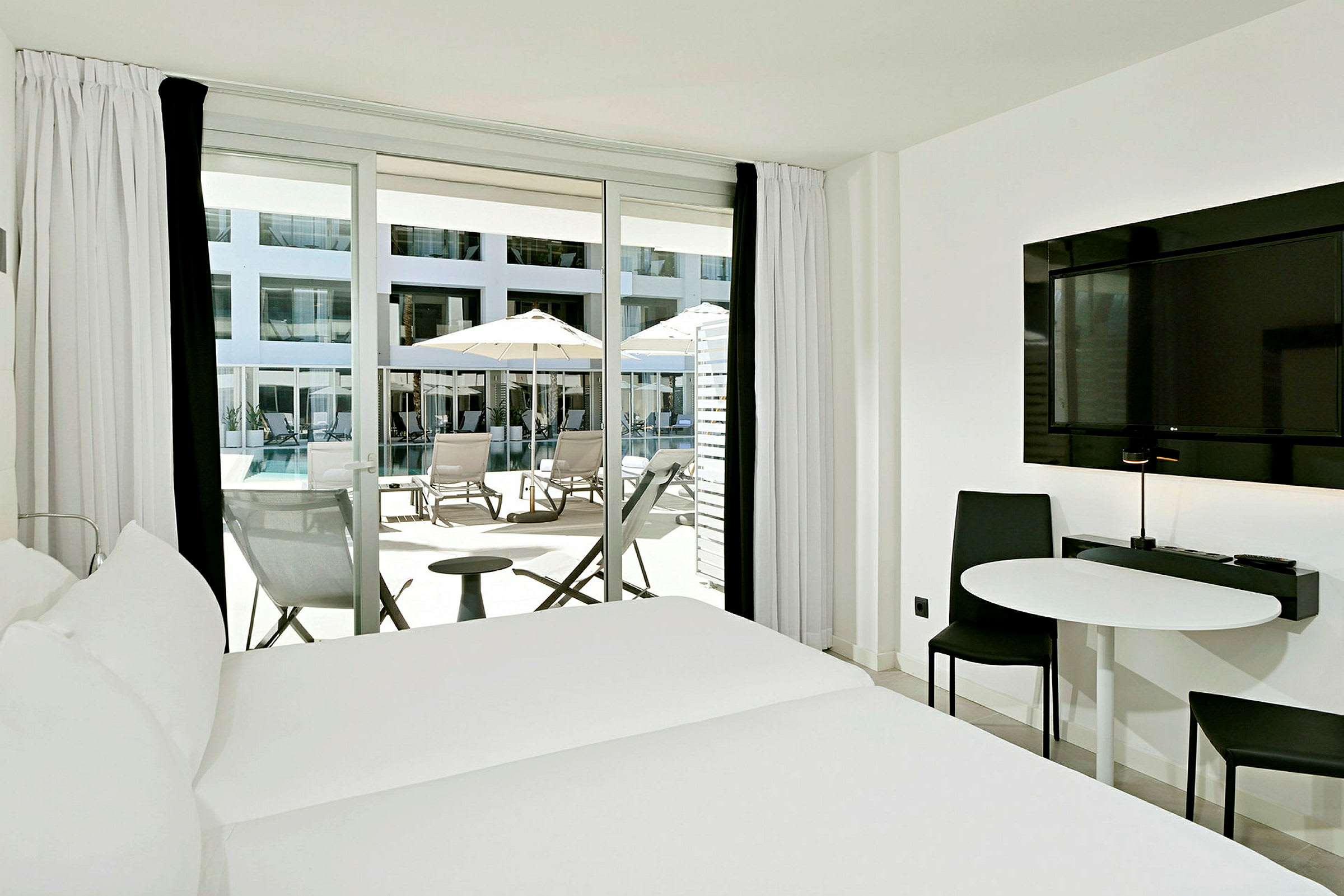 Hotellrum med säng, bord, stol och balkongdörr som leder ut till terrass med solstolar och parasoller vid en pool. (AI-genererad text)