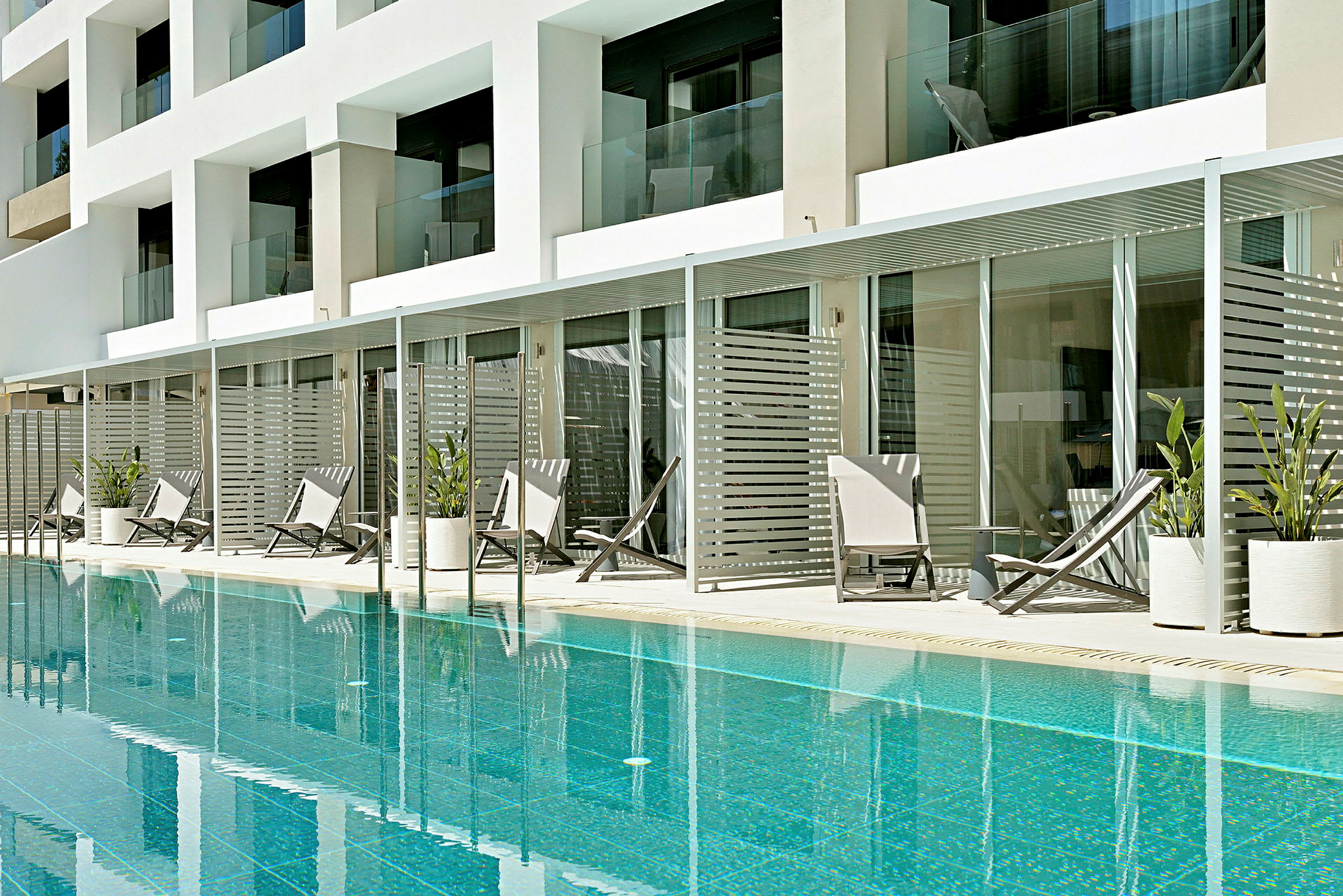 En swimmingpool framför en modern hotellbyggnad med stora balkonger och solstolar omgivna av planterade växter. (AI-genererad text)