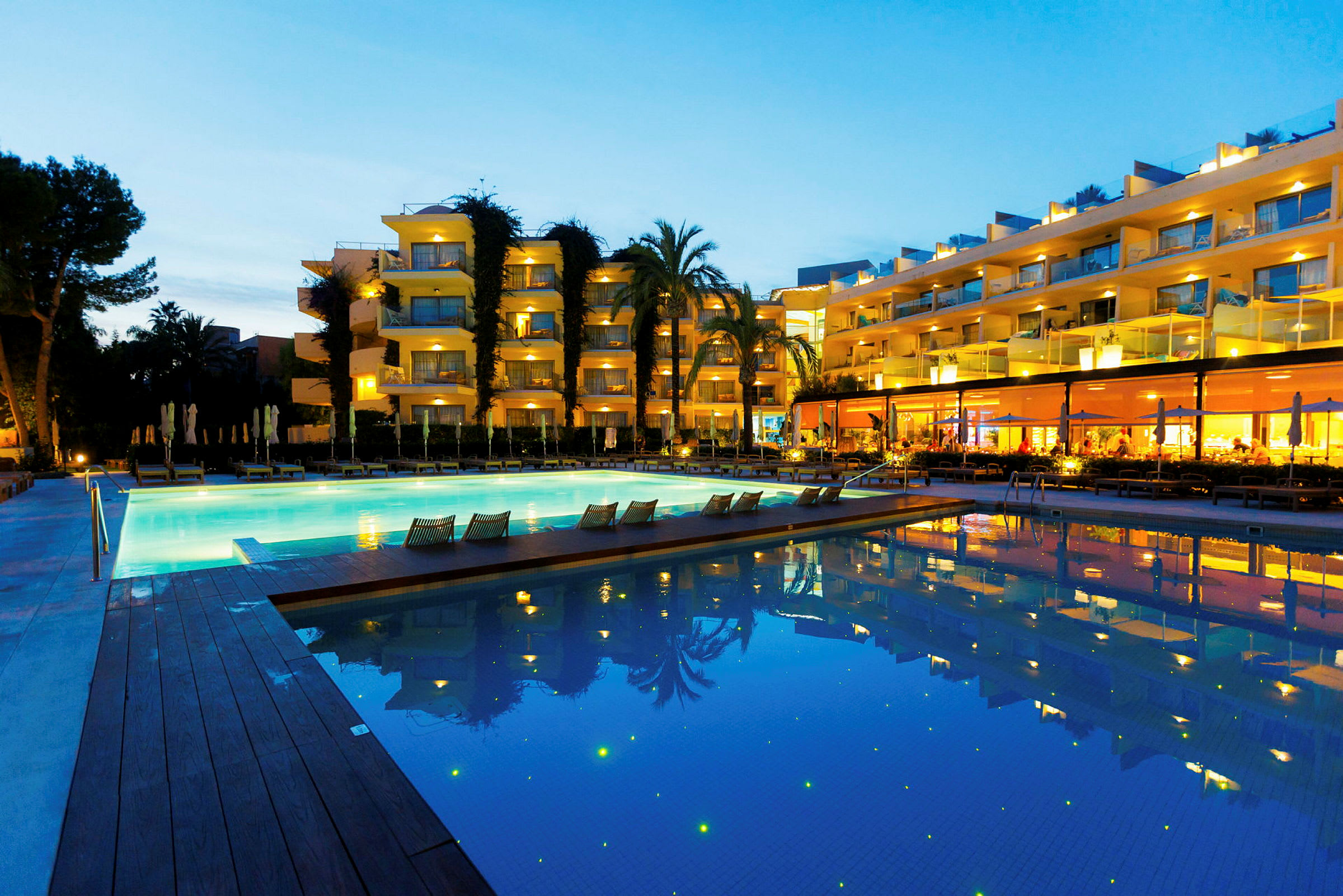 Hotell med ljusa fasader, en swimmingpool och omgivande grönska under kvällstid. (AI-genererad text)