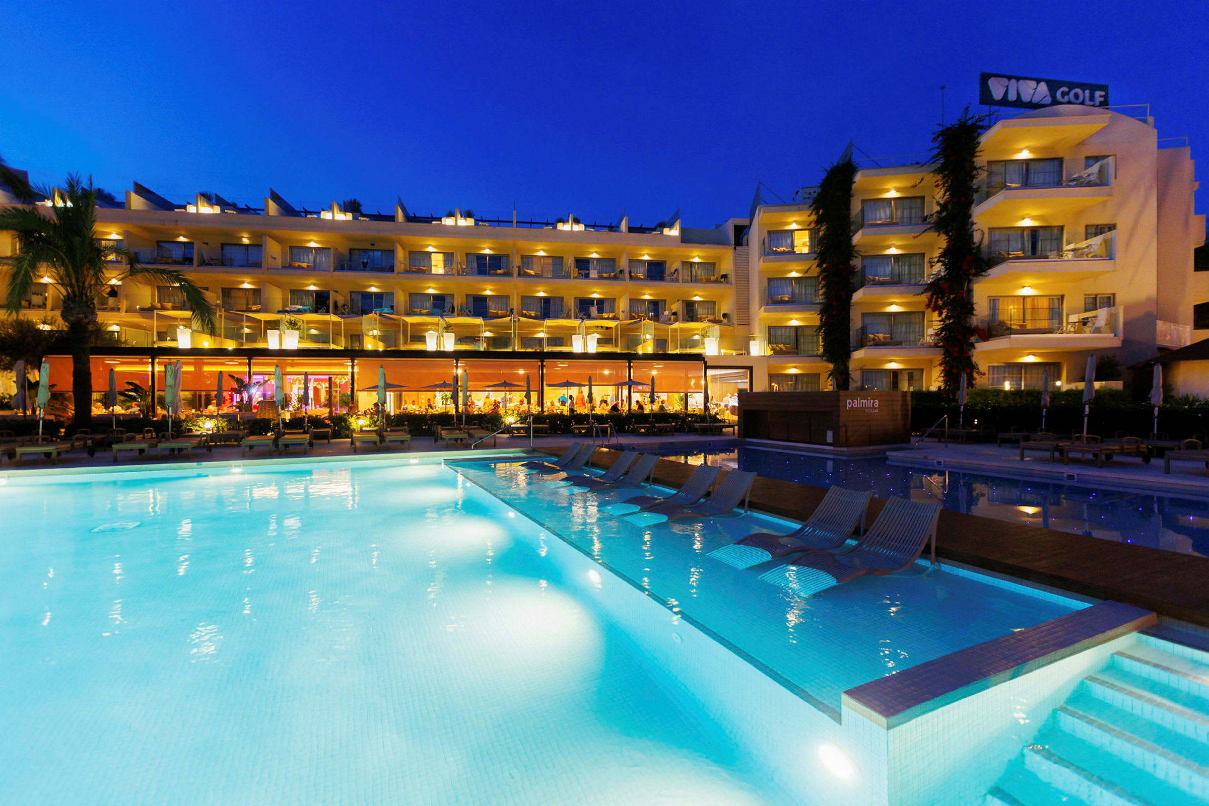 Hotellbyggnad med upplysta balkonger och en belyst pool i förgrunden på kvällen. (AI-genererad text)