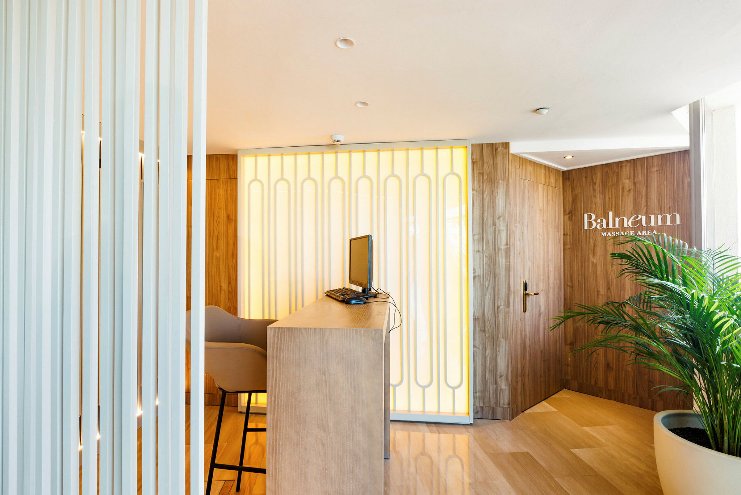 Reception med bord, dator och stol, med en bakgrund av väggpaneler och en dörr med identifiering 'Balneum Massage Area'. (AI-genererad text)