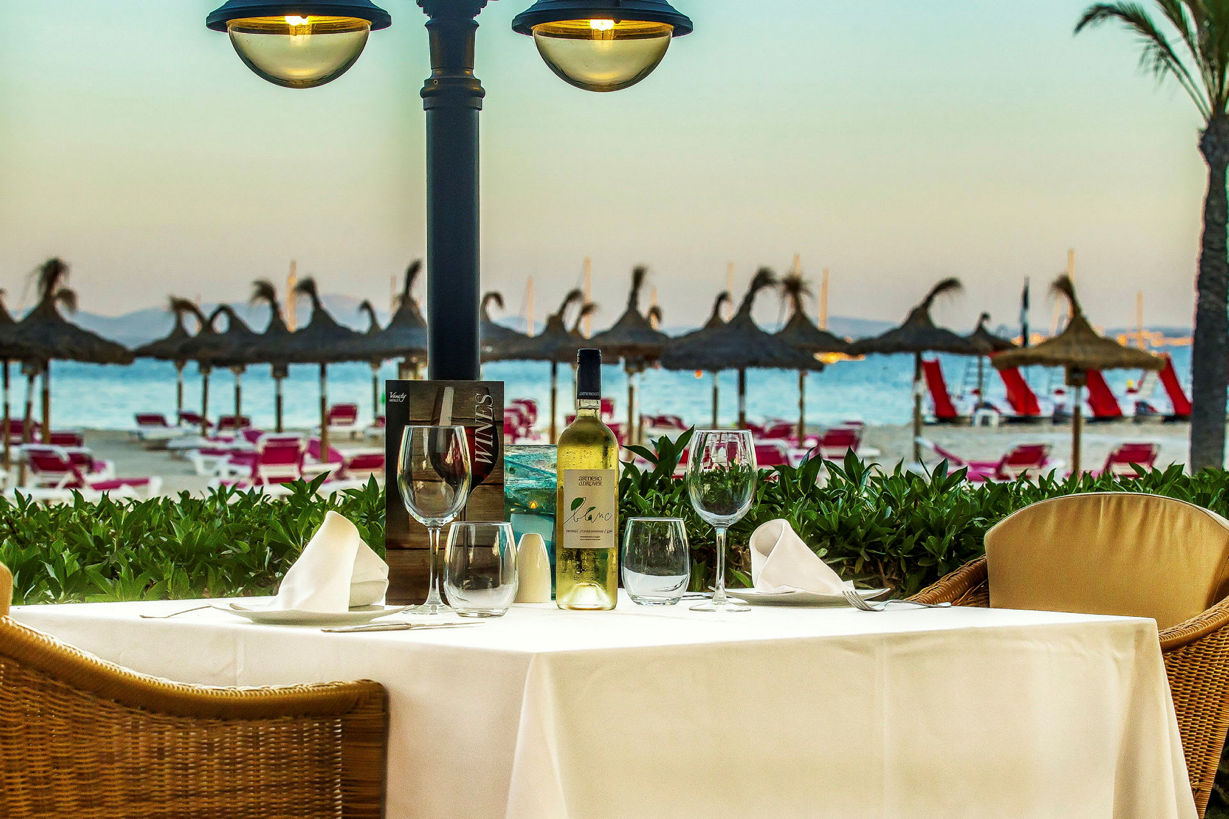 Ett bord med dukning och en vinflaska står på en terrass med utsikt över stranden och parasoller. (AI-genererad text)