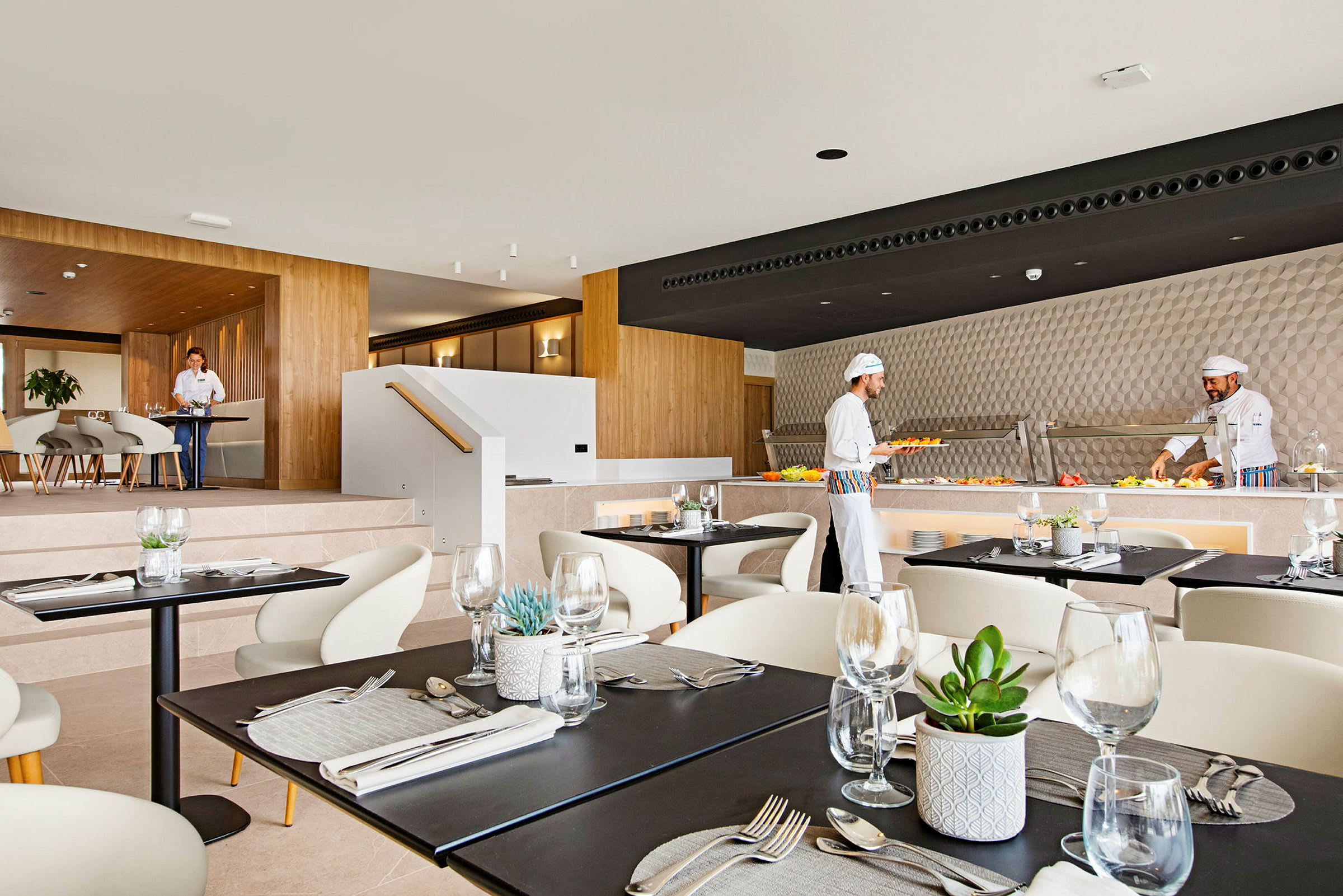 En modern restauranginteriör med dukade bord. Kockar förbereder mat vid en buffé. (AI-genererad text)