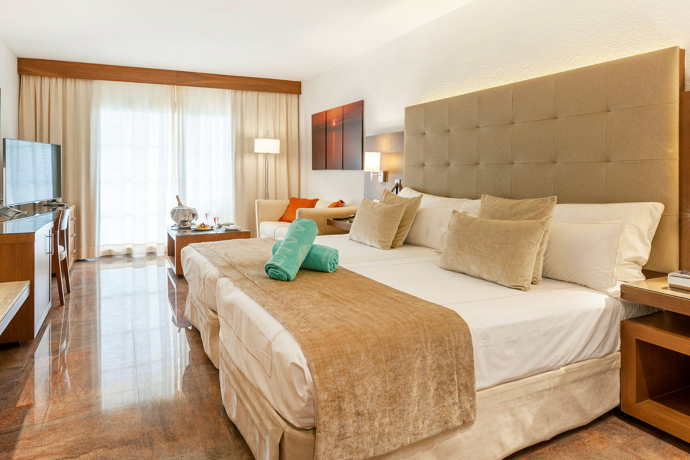 Hotellrum med en stor säng, sittgrupp och ljusa gardiner framför en balkong. (AI-genererad text)