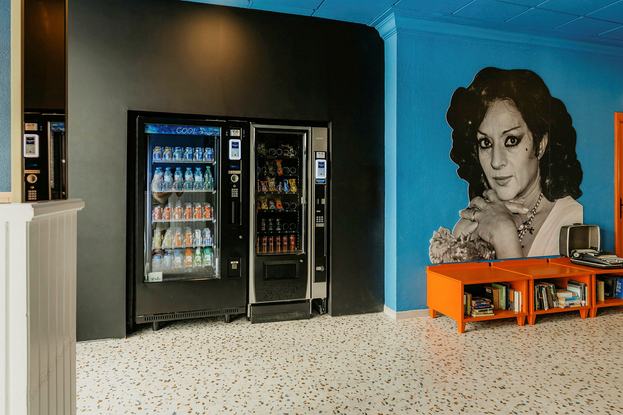 Bild på två automater för drycker och snacks placerade mot en svart vägg, tillsammans med ett område med orange hyllor innehållande böcker framför en blå vägg. (AI-genererad text)