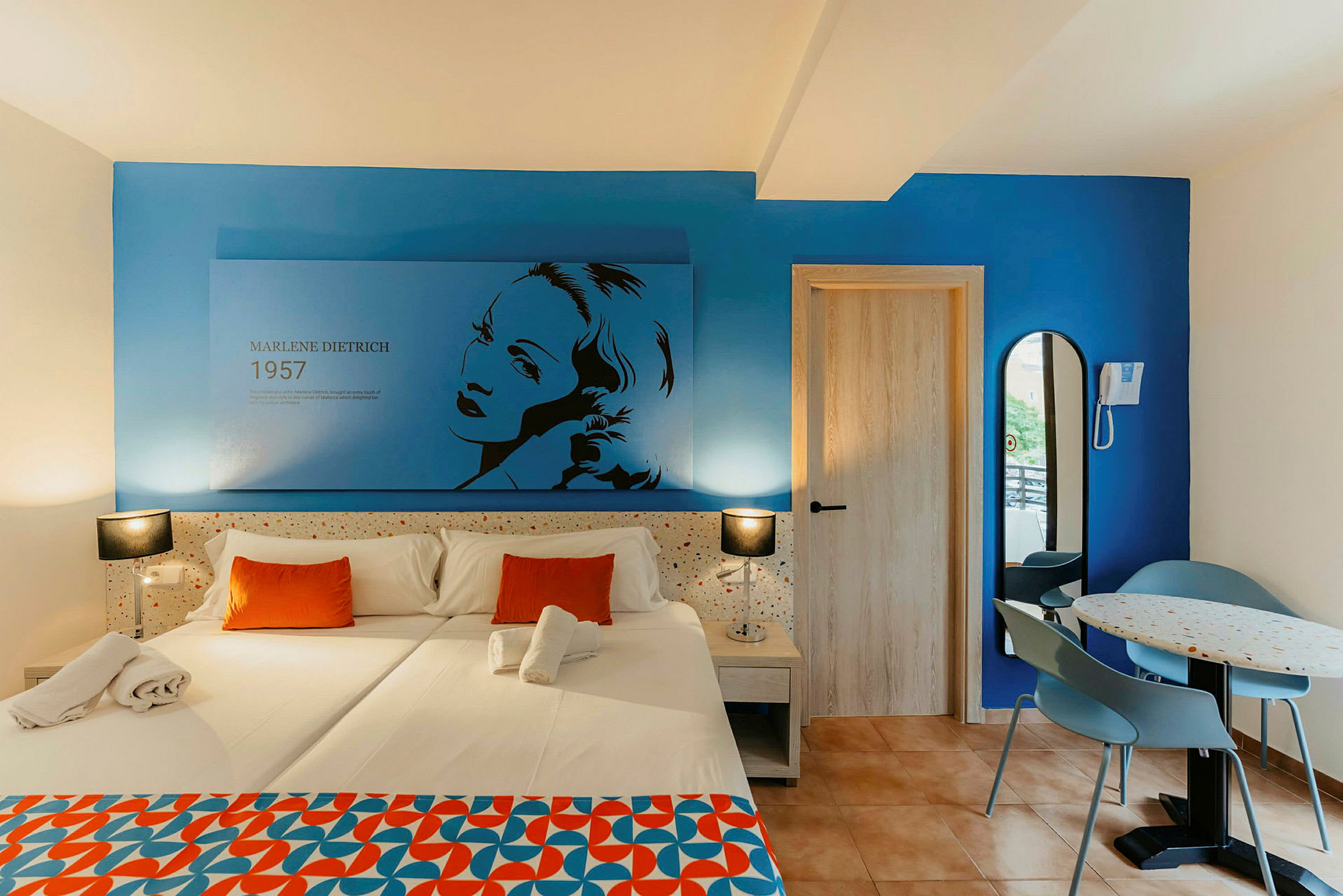 Ett hotellrum med en dubbelsäng klädd i vit linne och orange kuddar. Väggen är blå och har en illustration av Marlene Dietrich med text. Till höger finns en dörr och en liten matplats med ett bord och två stolar. (AI-genererad text)
