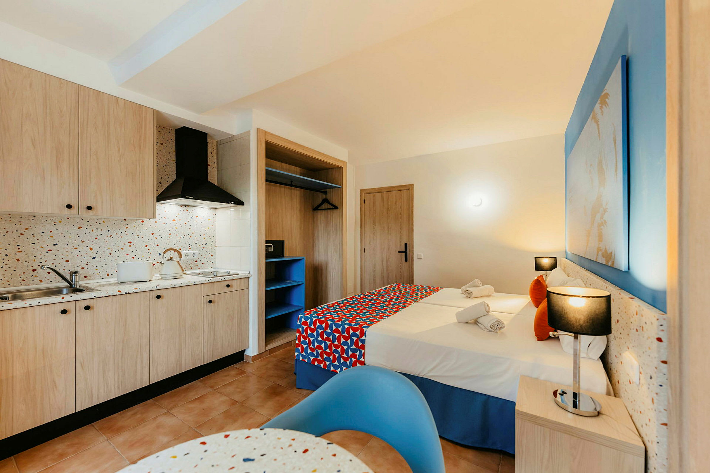 Ett hotellrum med kök och sovområde. Köket har träskåp, diskbänk och spis. Sovområdet har två bäddar med färgglada överkast samt sängbord och belysning. (AI-genererad text)