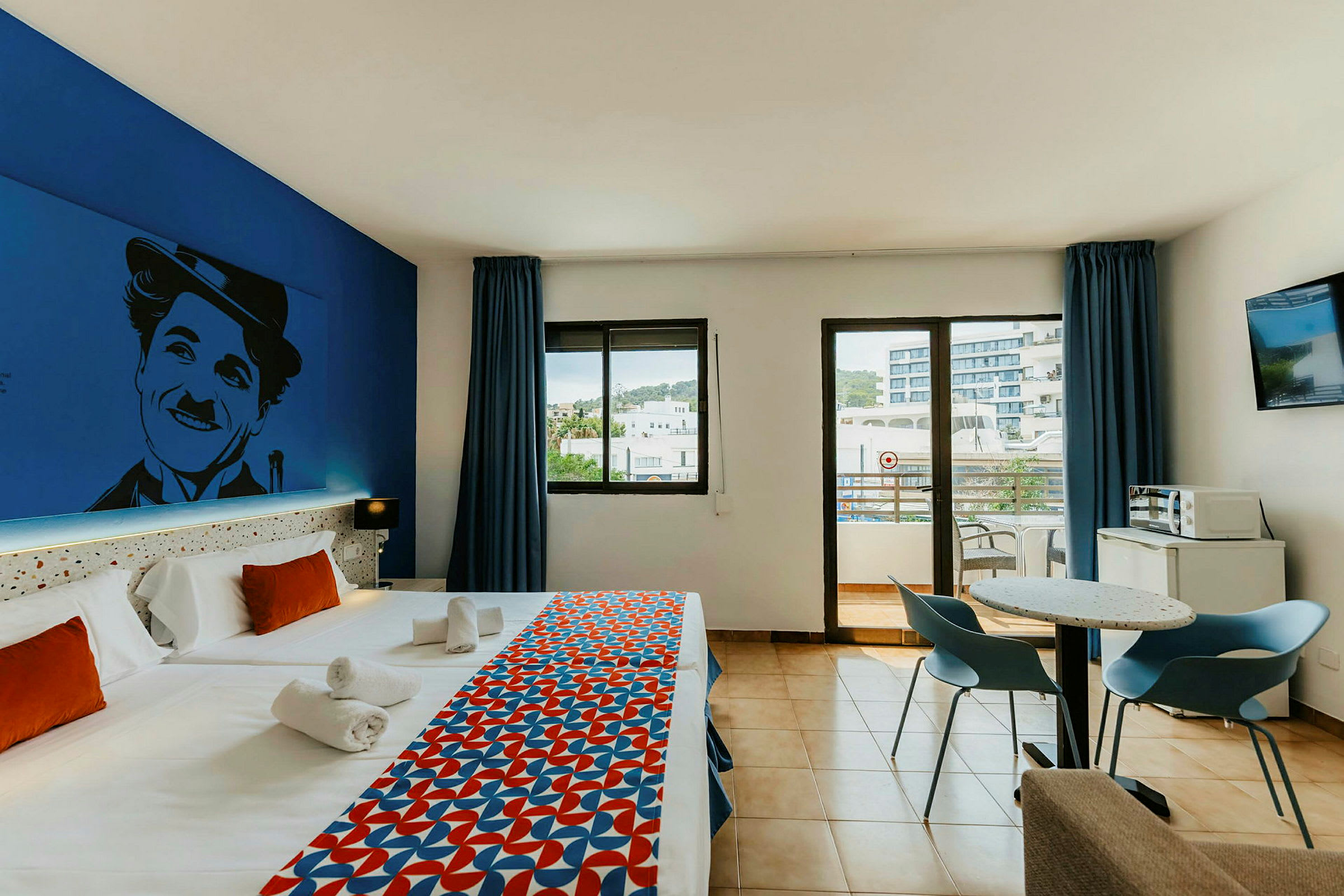 Hotellrum med två enkelsängar, färgglad sängduk, väggdekoration, bord och stolar, samt utsikt mot en balkong. (AI-genererad text)