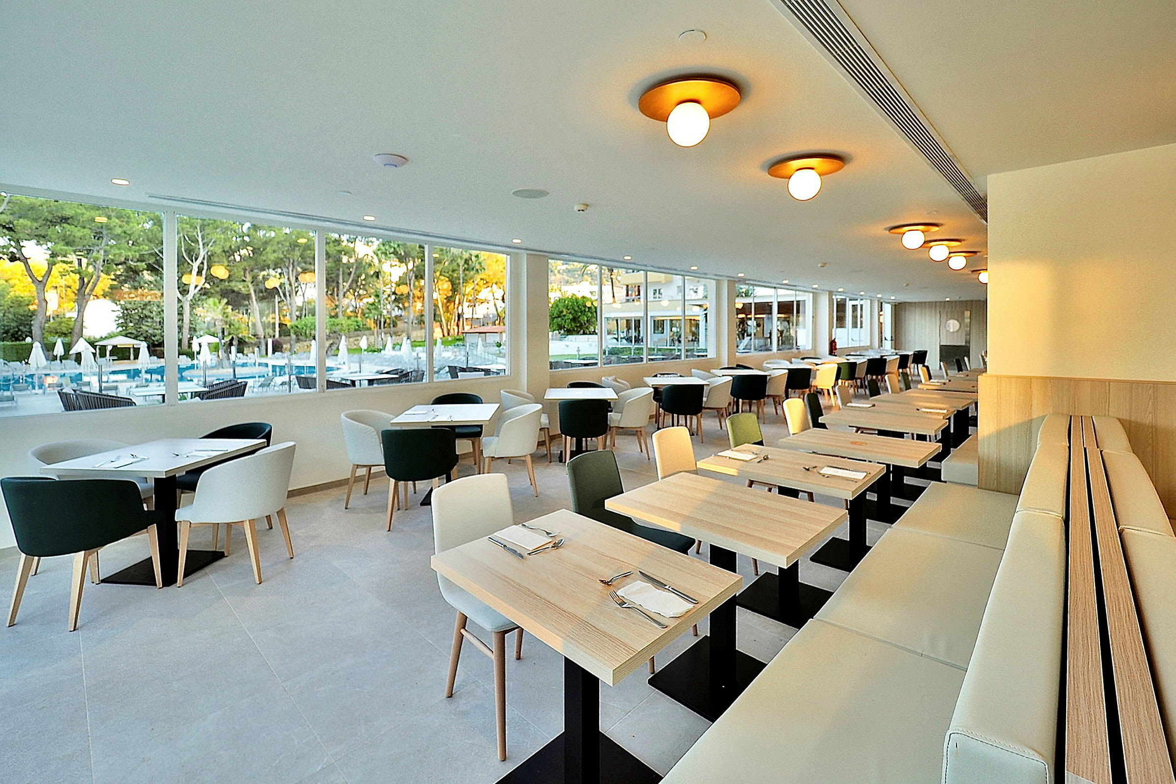 En modern restaurang med fönster som vetter mot en utomhuspool och grönska. Bord och stolar är ljusa och enkelt designade. (AI-genererad text)