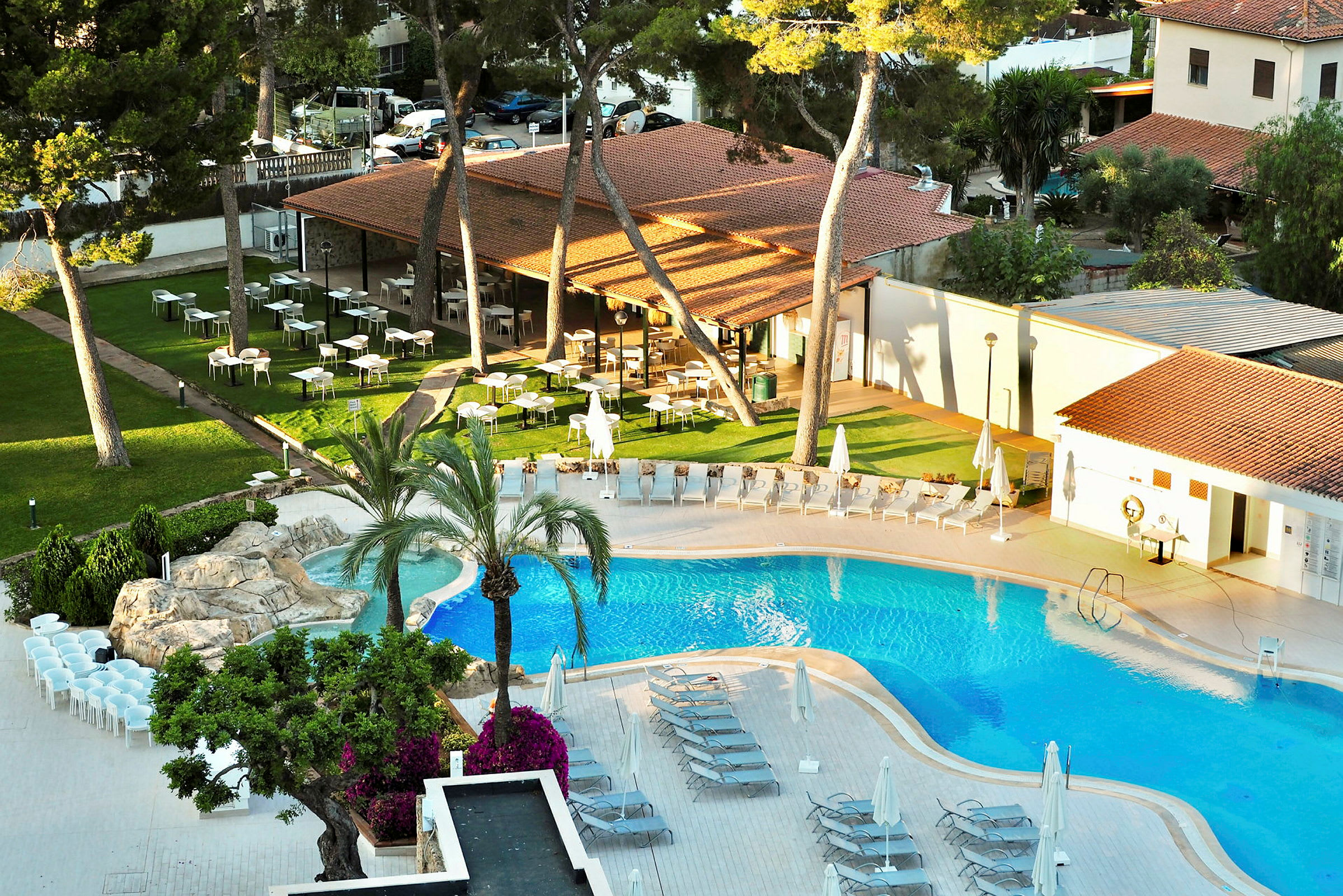 En bild av en pool som omges av solstolar och parasoller. I bakgrunden finns en gräsmatta med bord och stolar samt en byggnad med tegeltak. (AI-genererad text)